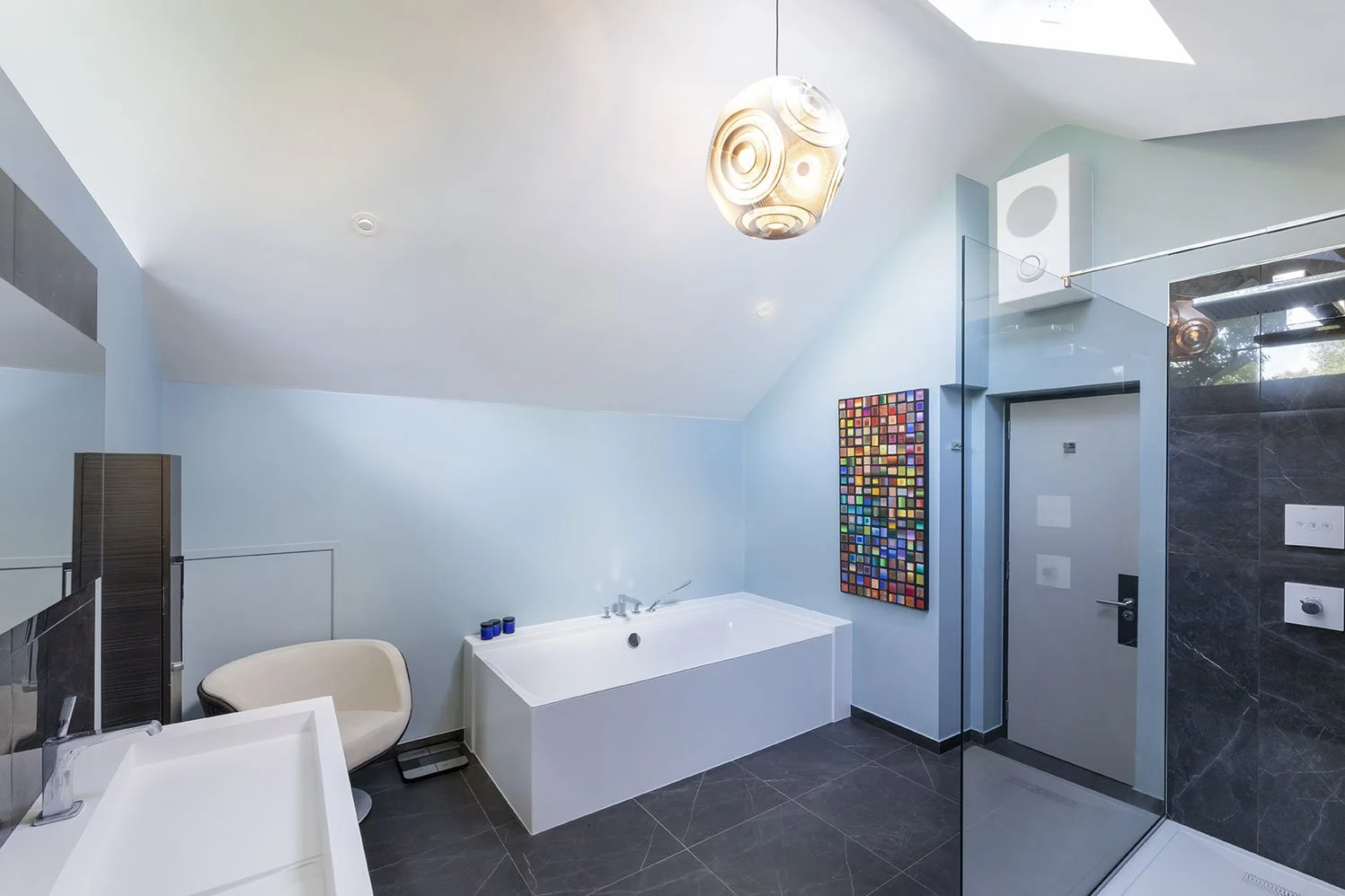 Modern bathroom with a white bathtub, a beige chair, colorful wall art, and a glass shower enclosure. The room has light blue walls and dark gray tiled floors.