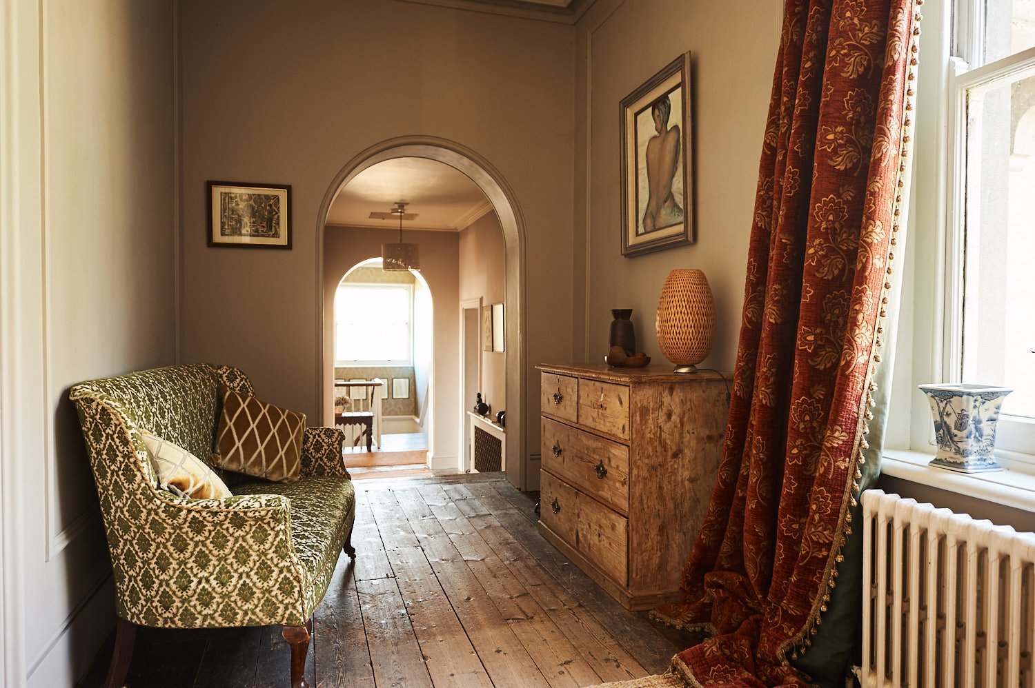 Living room with wooden floor, vintage style sofa, framed artwork on the wall, wooden chest of drawers with vases, patterned curtains, large window, and hallway leading to another room.