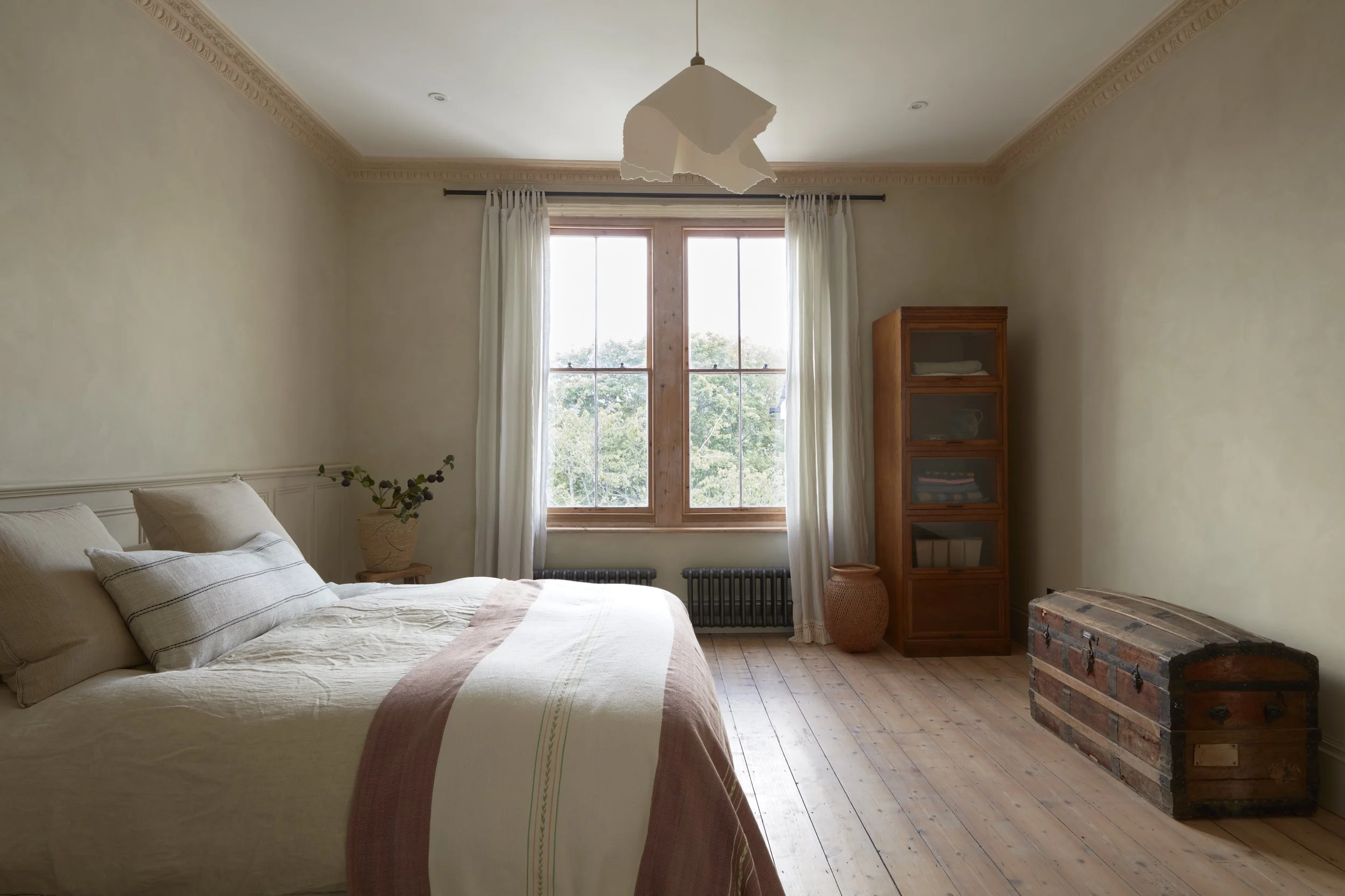 A cozy bedroom with a large window, cream-colored walls, a bed with neutral bedding, and wooden furniture including a chest and a cabinet.