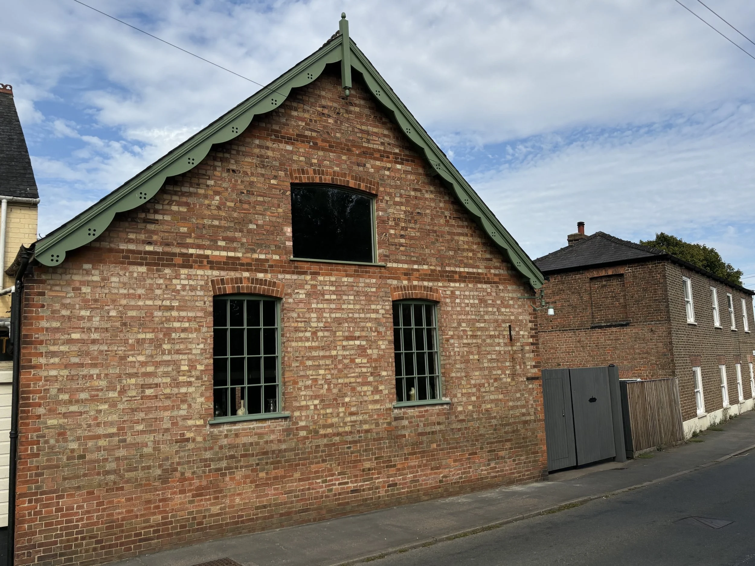 A brick house with a green gabled roof and three visible windows, two with bars and one without, situated along a paved street under a partly cloudy sky.