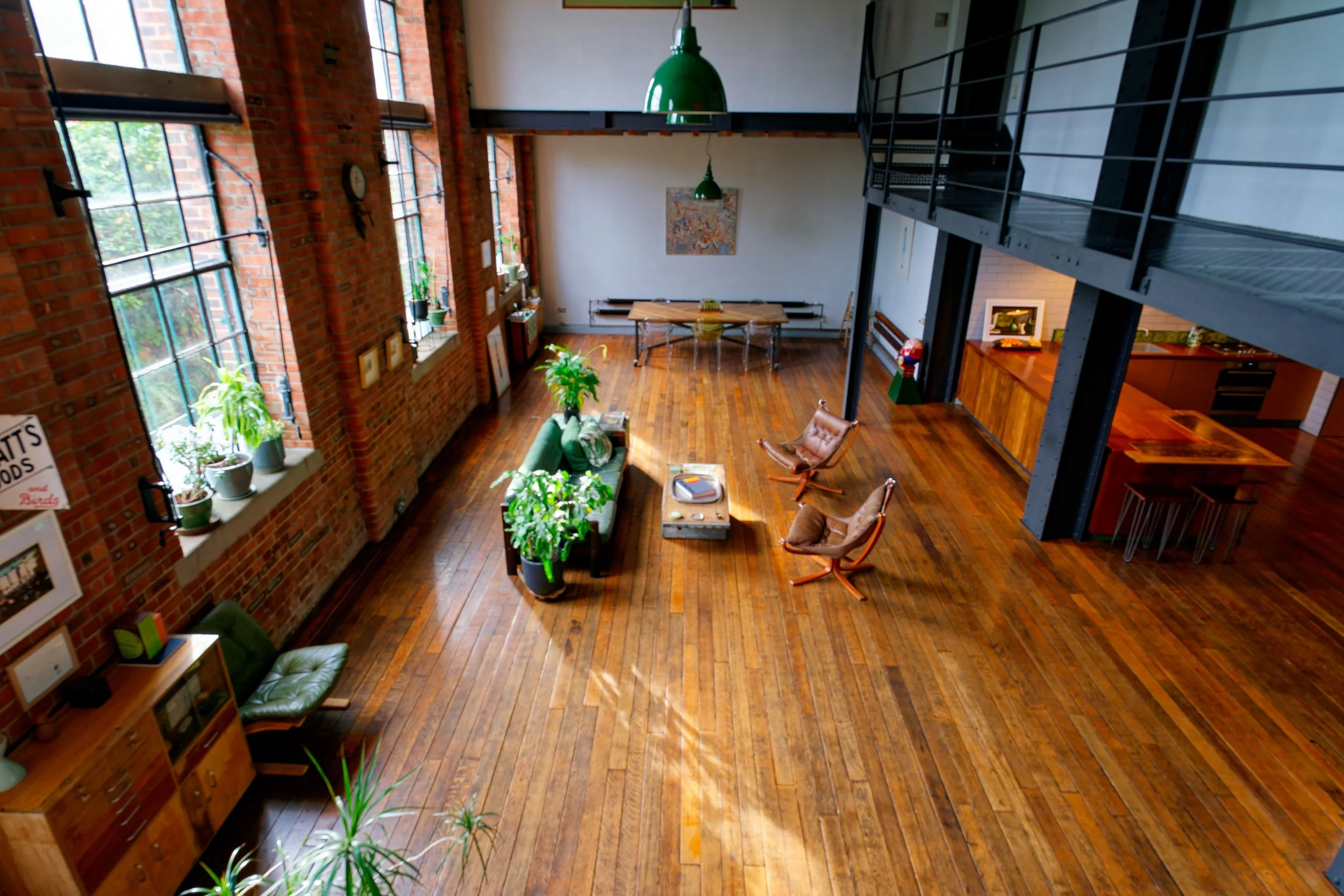 Spacious loft-style living area with exposed brick walls, large windows with potted plants, and wooden floors. Contains a green sofa, small table, lounge chairs, and wall art. Modern industrial open-plan design with upstairs railing and a small kitch