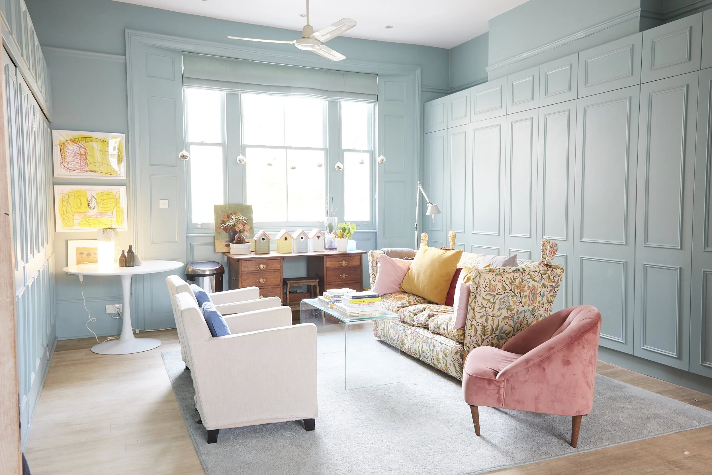 Bright living room with light blue walls and large windows. Contains a floral sofa, pink velvet chair, white armchairs, a wooden desk, and a small white table with decorative items.