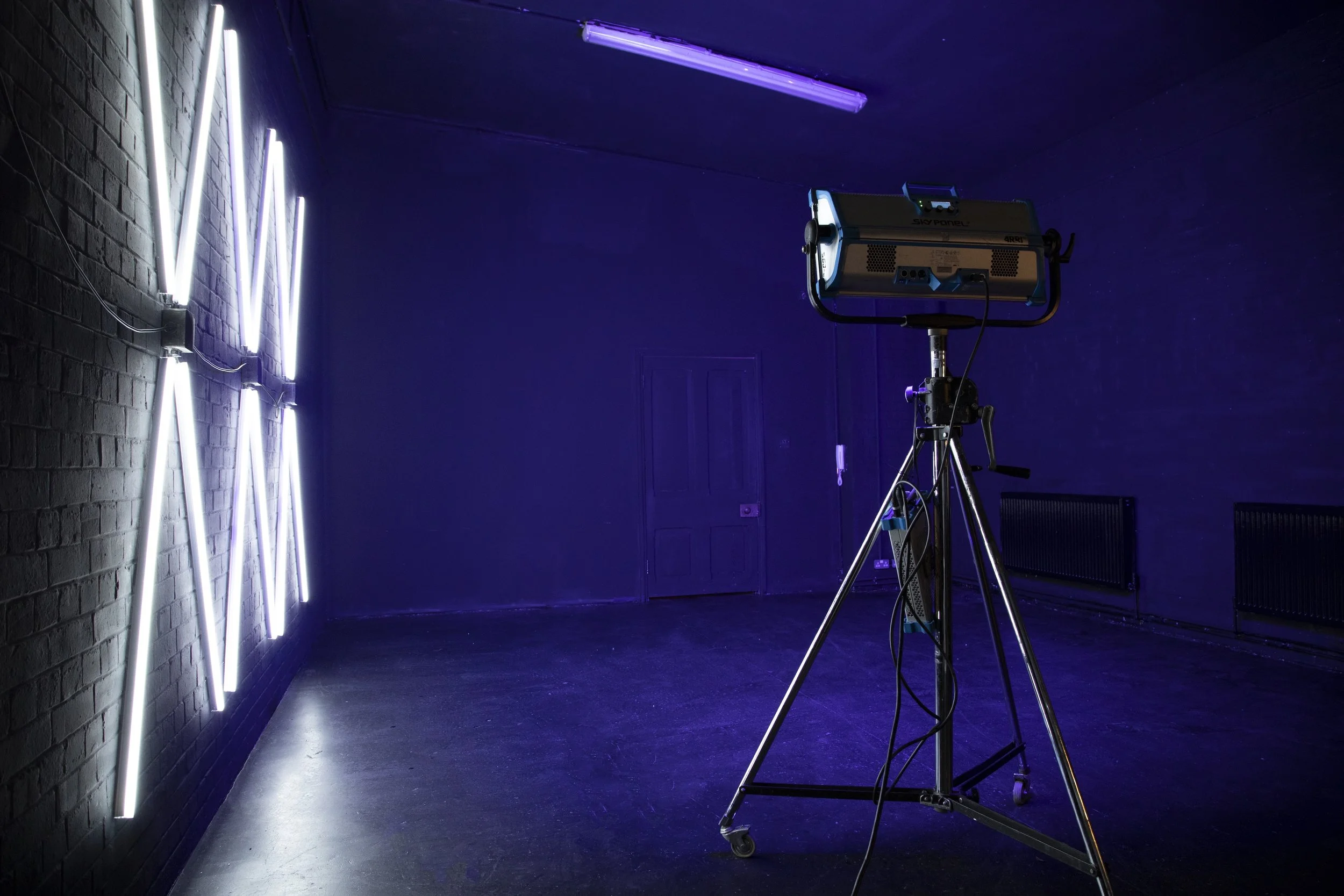 An empty art gallery with illuminated LED light art on a dark brick wall, and a large professional studio light on a tripod in the room.