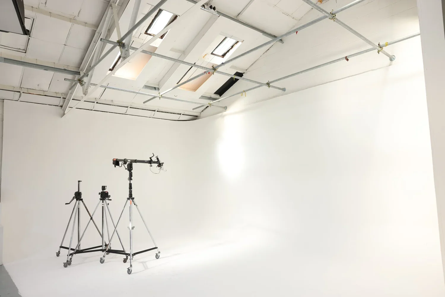 A white photography studio with three tripods and camera equipment set up on a white floor, and ceiling windows providing natural lighting.