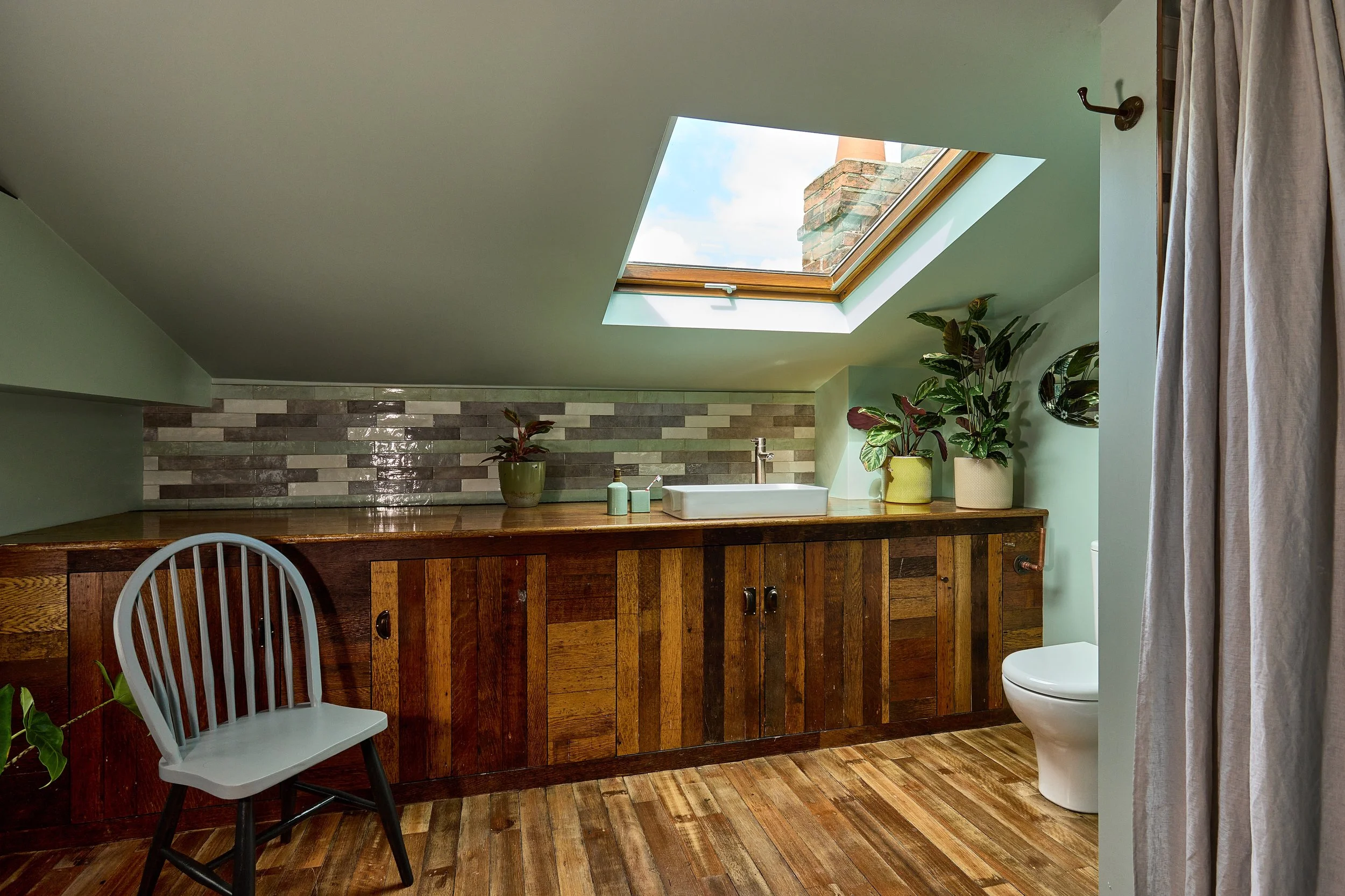 Bathroom with sloped ceiling, skylight window, wood cabinetry, potted plants, toilet, and partially visible curtain.