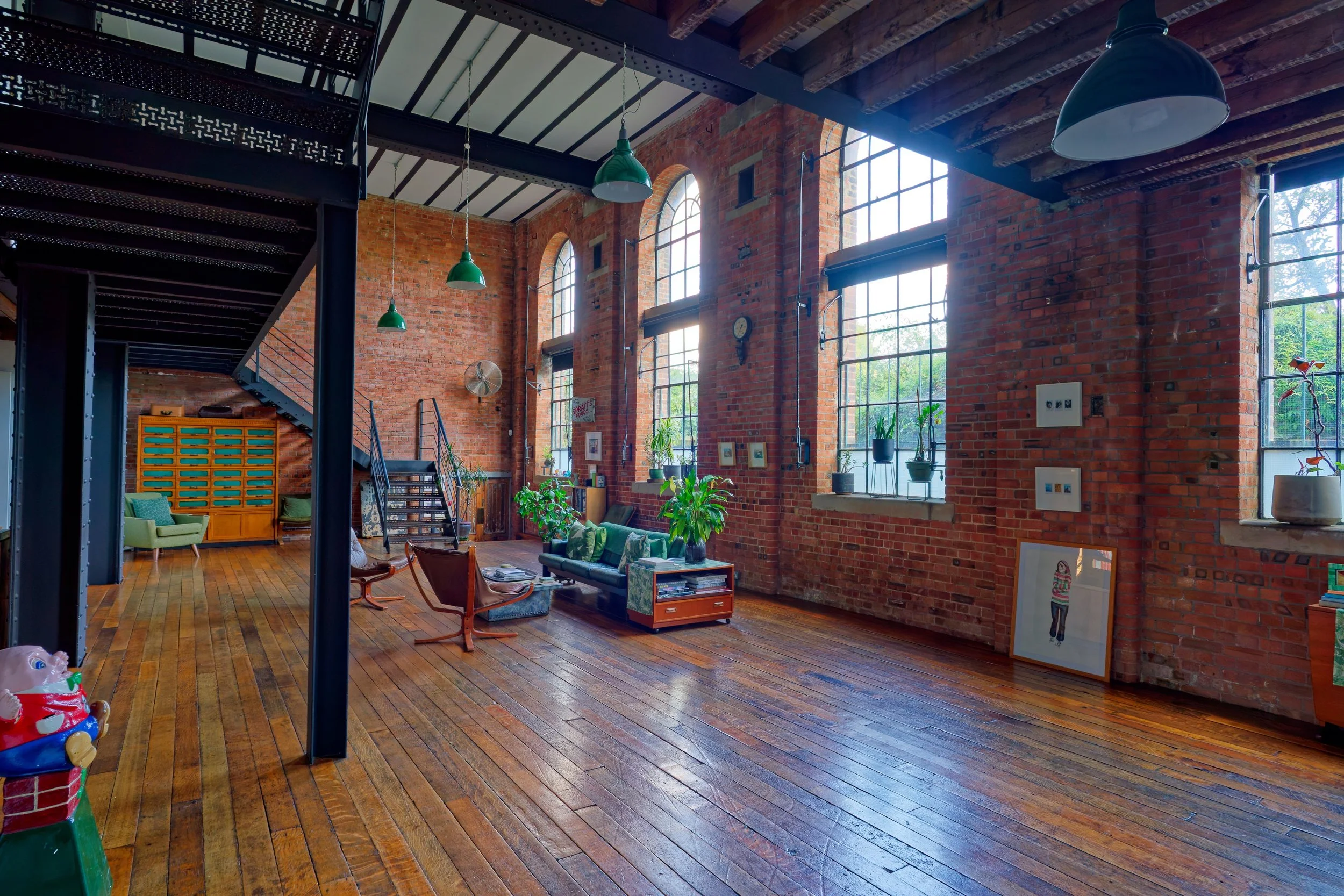 Spacious loft with exposed brick walls, large industrial windows, green pendant lights, and wooden floor. Contains seating area with a sofa, armchairs, and shelves with plants and art decor.