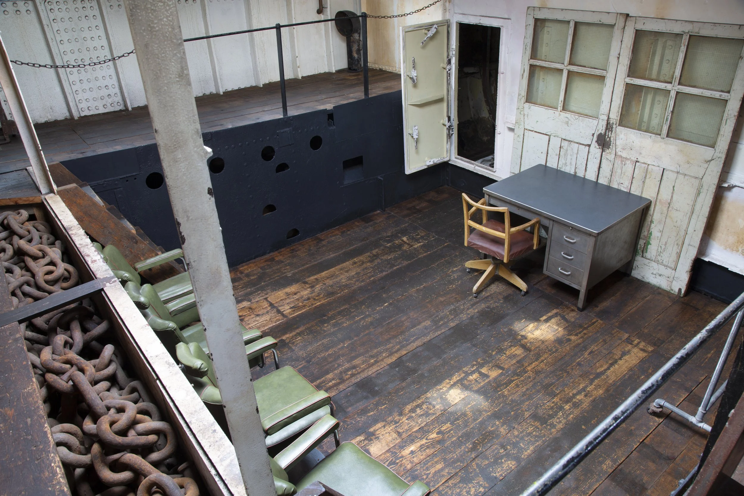 Empty industrial-style room with a wooden floor, an old desk with a chair, metal cabinets, and a row of green chairs along a railing with rusty chains.