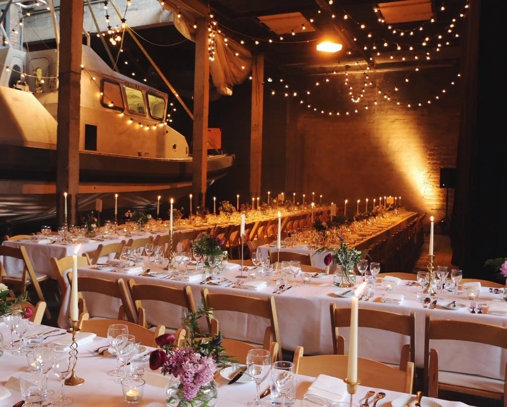 Long banquet tables set up for a celebration or wedding in a rustic venue with string lights, candles, floral arrangements, and a vintage boat displayed on a second level.