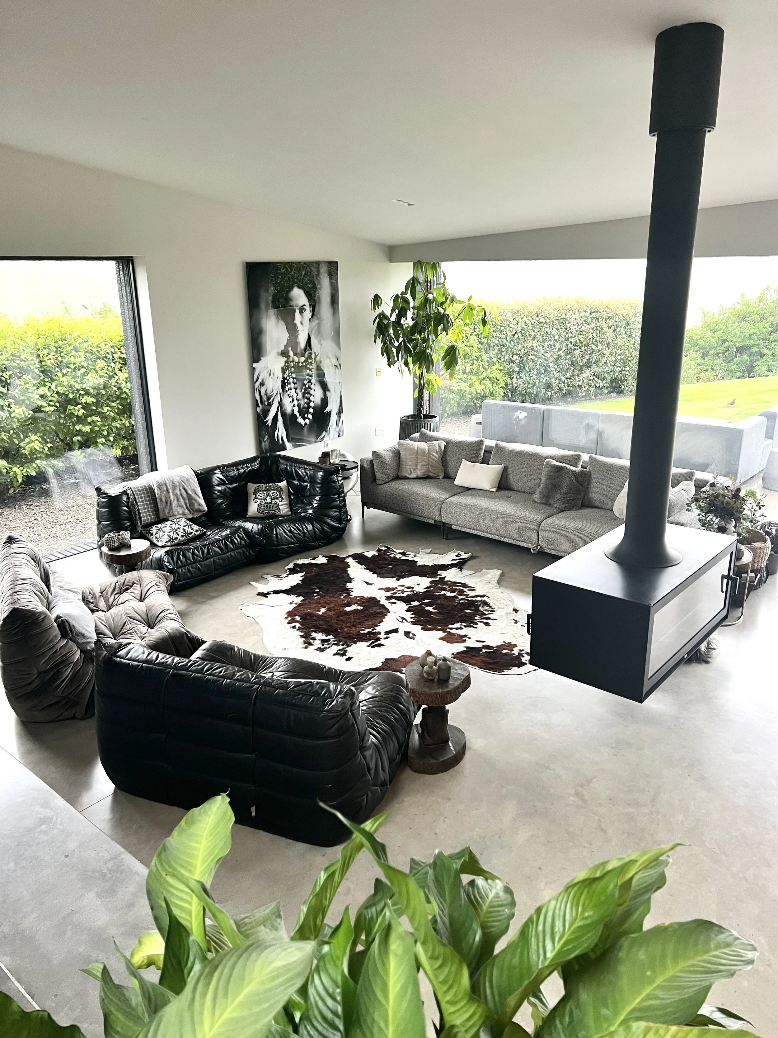 Modern living room with large windows, black and gray sofas, a cowhide rug, a black and white portrait on the wall, and greenery visible outside.