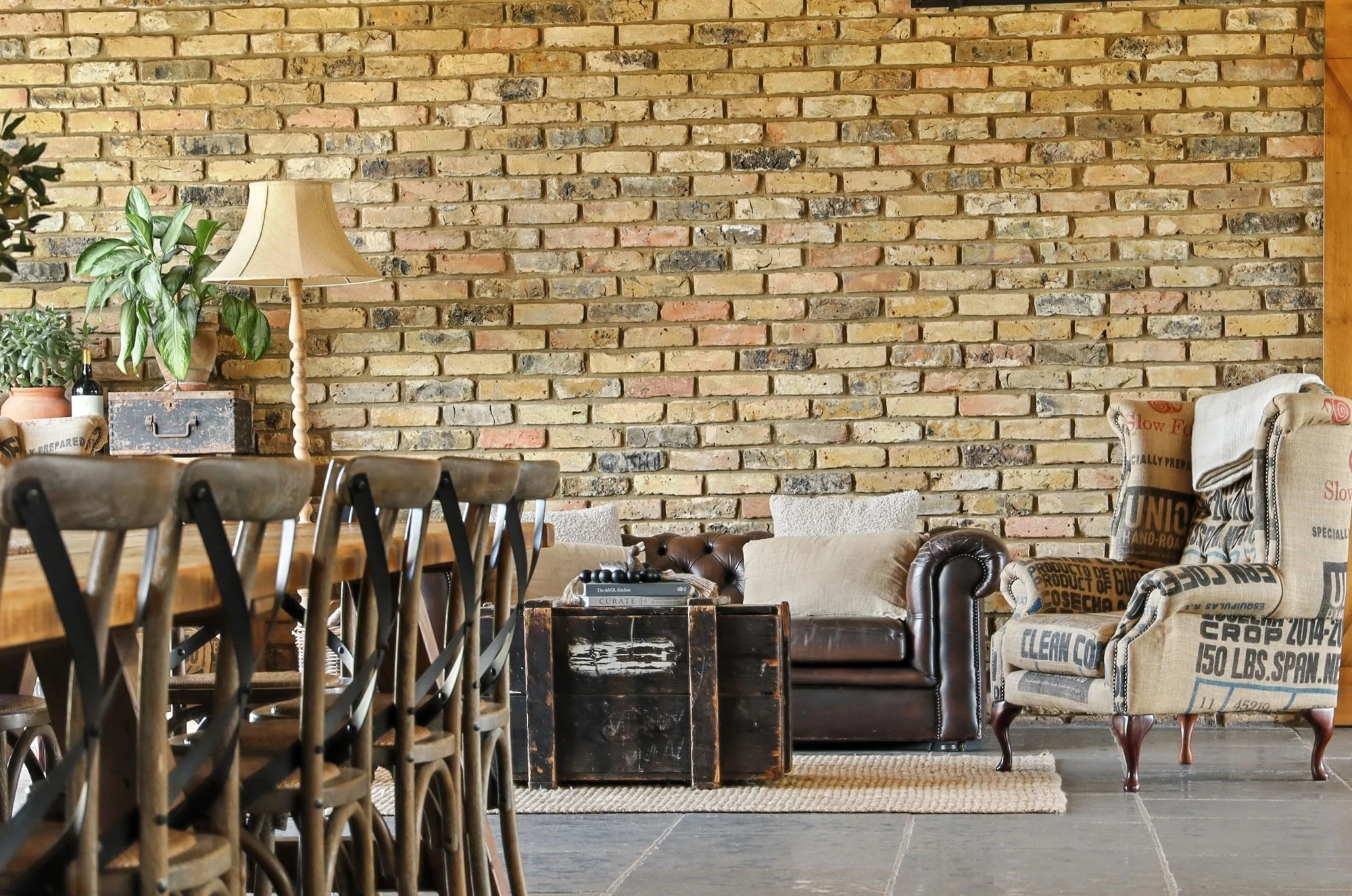 A cozy living room with a brick wall background, featuring a beige armchair with printed fabric, a dark brown leather armchair, a wooden table, and potted plants, including a leafy green plant and a smaller succulent. A standing lamp with a beige sha