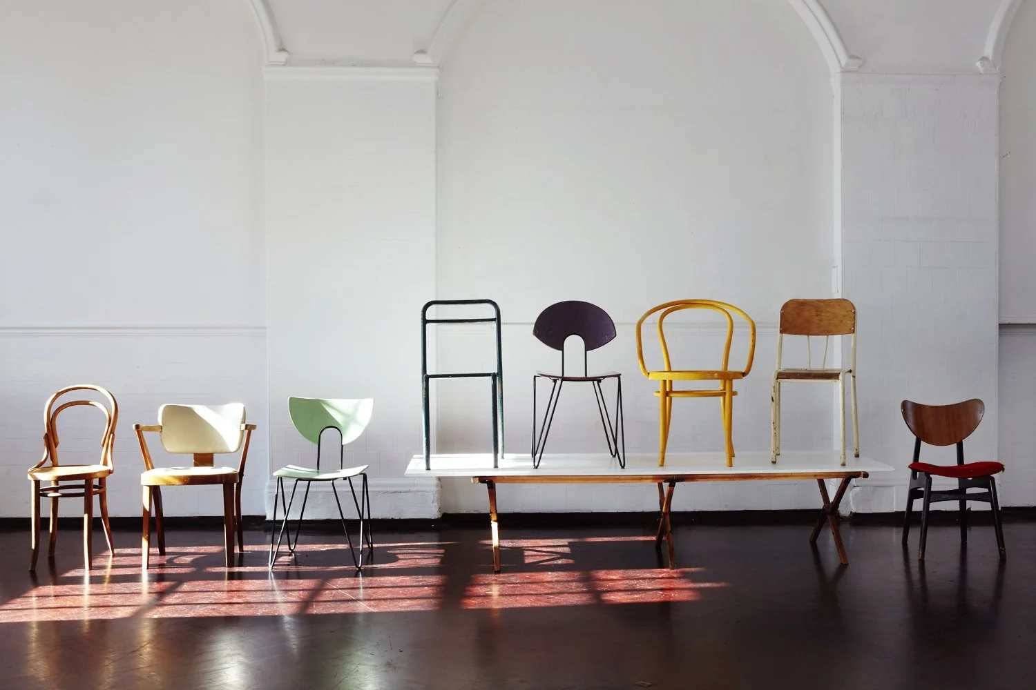 A collection of colorful chairs arranged in a line on a platform against a white wall with sunlight casting shadows on the dark wooden floor.