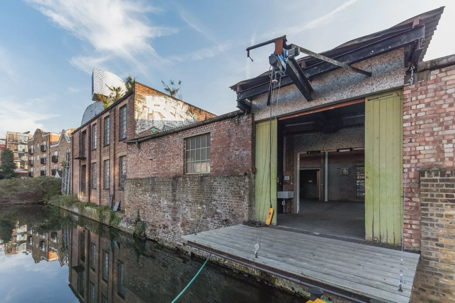 Old brick building next to a canal with a platform extending over the water, some construction or renovation equipment on the platform, and an open doorway leading inside.