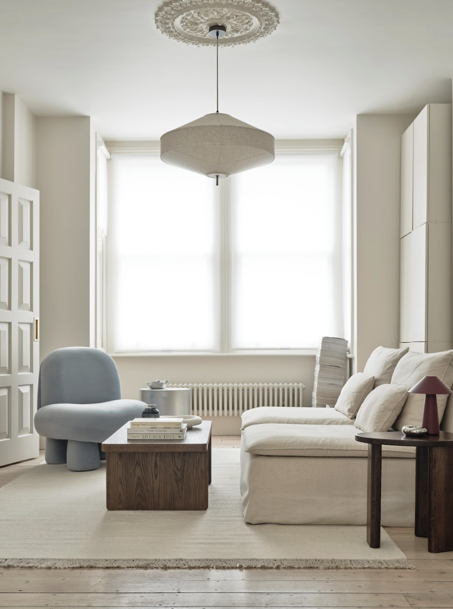 A bright, minimalist living room with large windows, a white sofa, a light blue lounge chair, a wooden coffee table with books, and a side table with a red lamp. The room features neutral tones and a modern design.