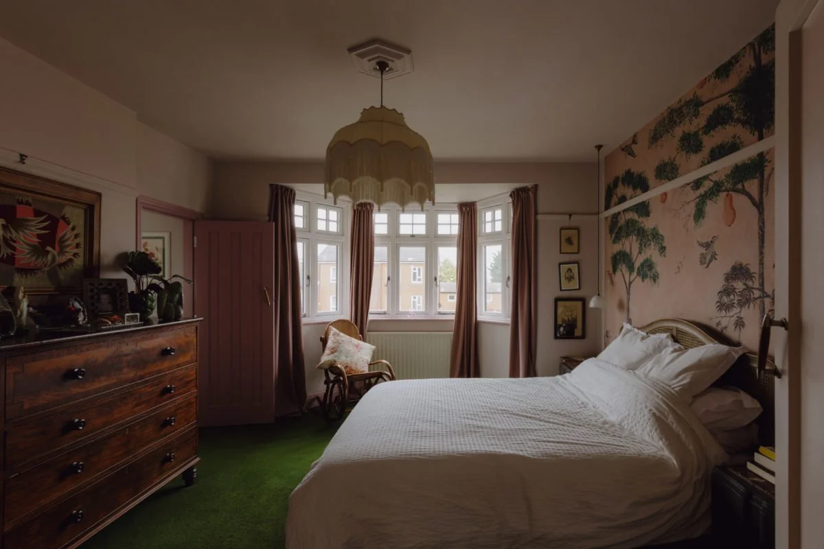 Cozy bedroom with a vintage vibe, featuring a large bed with white bedding, an ornate headboard, a wooden dresser with framed photos and decorations, a rocking chair with a pillow near a bay window, mustard curtains, and a floral wallpaper mural on o