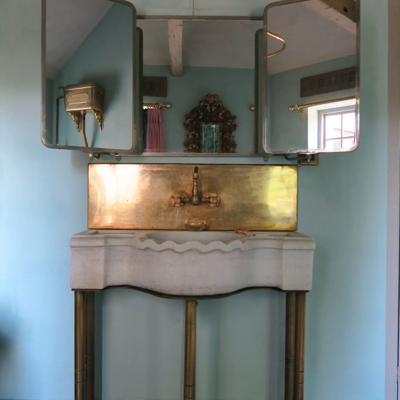 Vintage bathroom sink with a white marble basin and brass fixtures, mounted on four brass legs, with an antique gold mirror above reflecting a room with a window and decorative items.