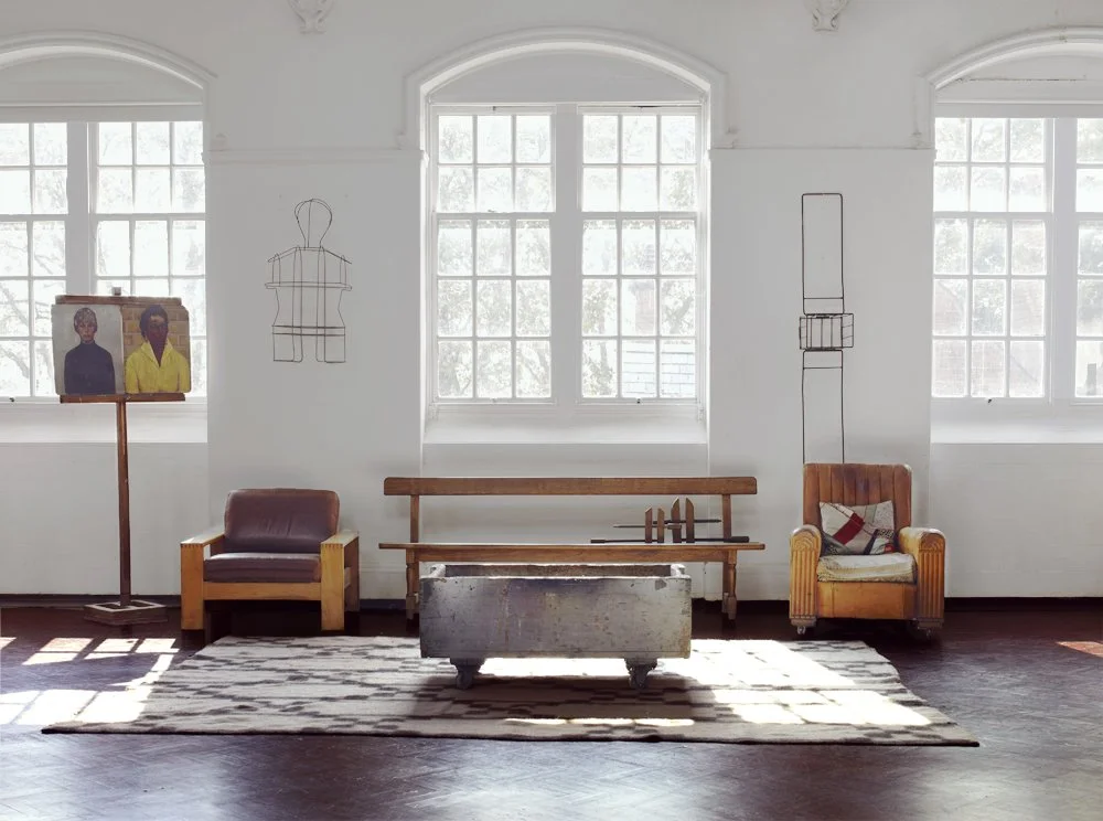 Minimalist living room with three large windows, a wooden bench, two armchairs, a coffee table, and wall art featuring abstract outlines of figures and a painting of two people, all in a bright, airy space.