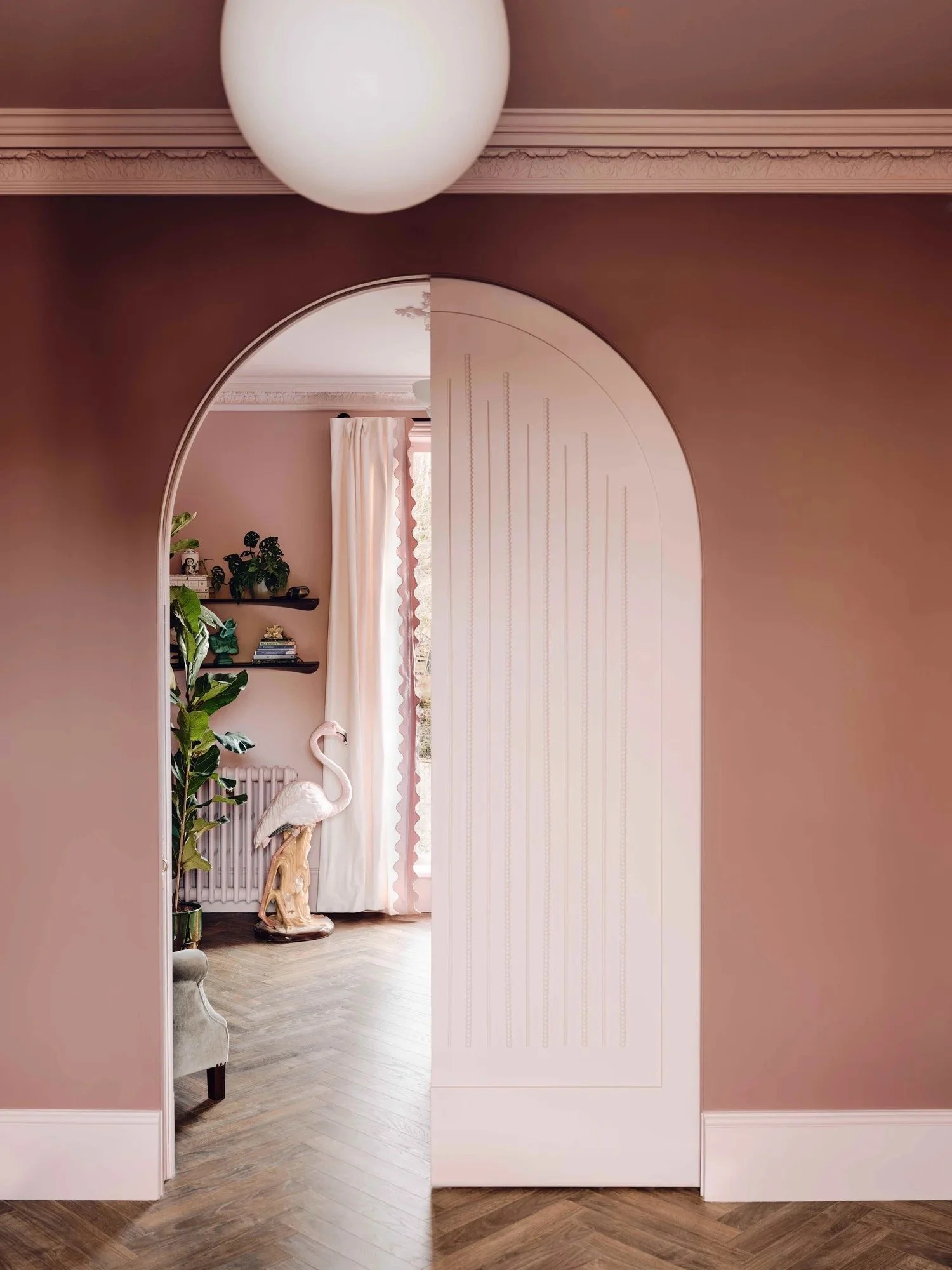 Interior view of a room seen through an arched doorway, featuring pink walls, a white curtain, and a decorative flamingo statue beside a potted plant and wall shelves.