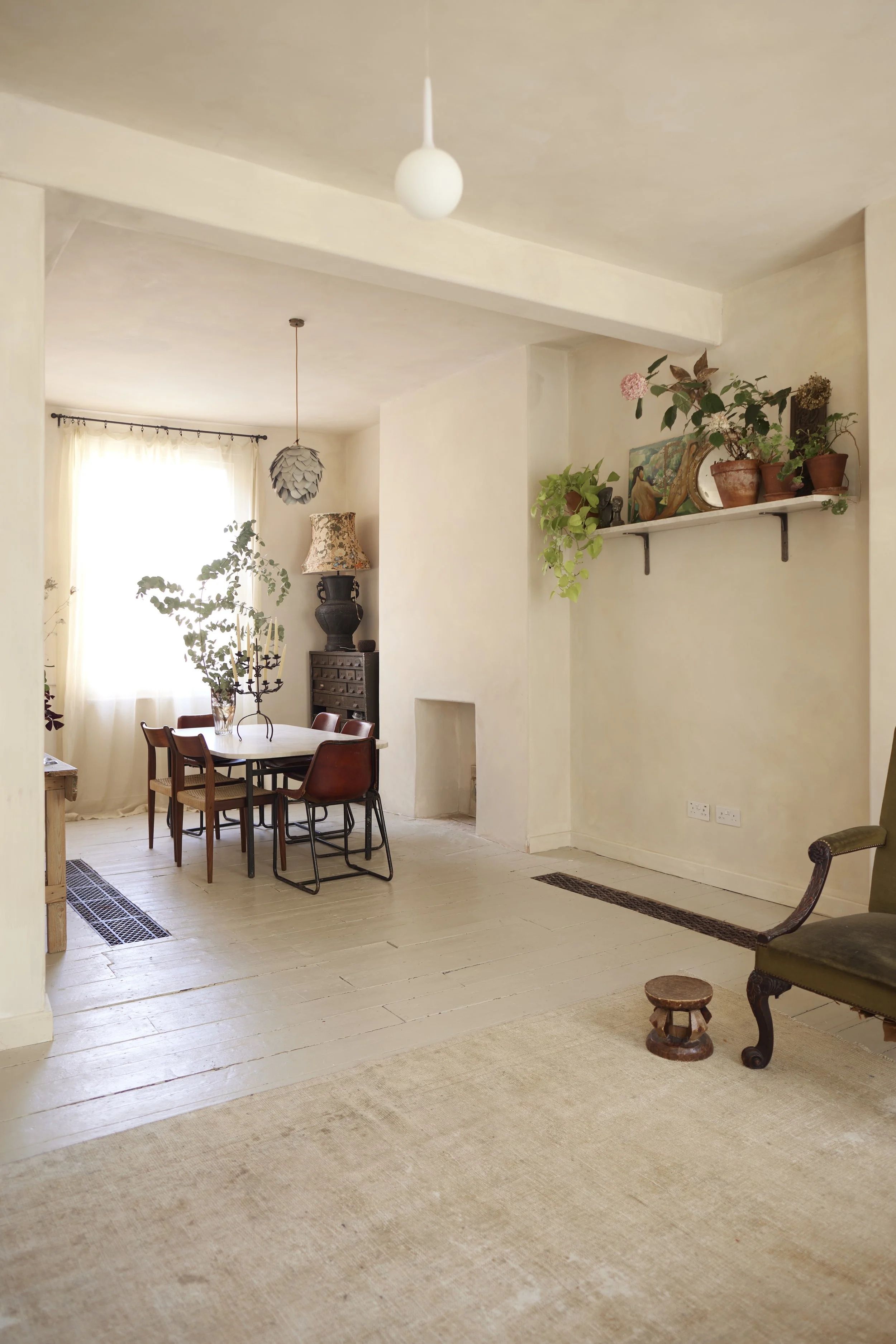 A cozy dining area with a table and vintage chairs, a window with sheer curtains, and a shelf with potted plants and decorative items, all illuminated by natural light.