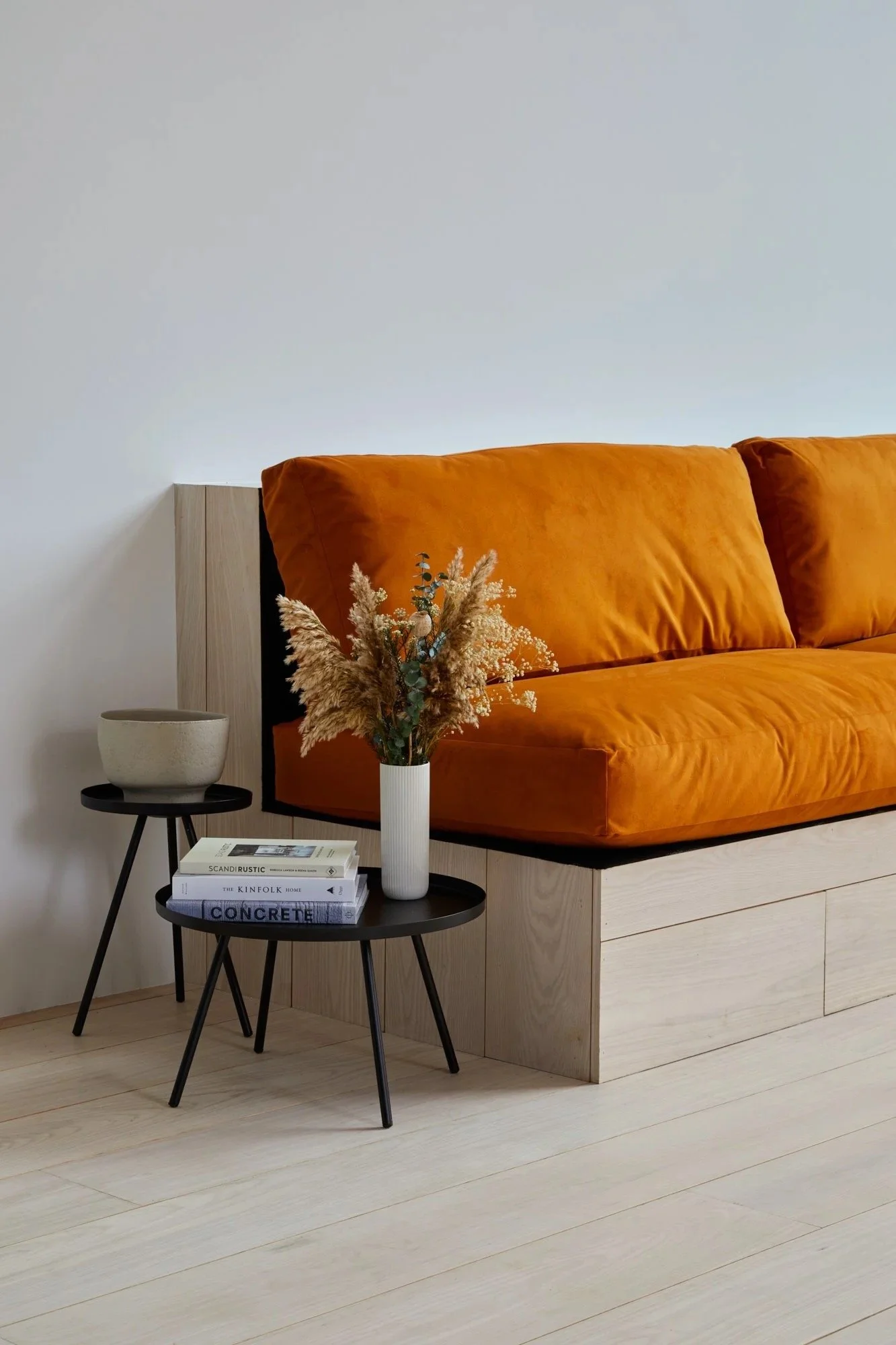 Living room corner with an orange velvet sofa, two black round side tables, a white vase with dried flowers, and a stack of books on the lower table, against a plain white wall and light wooden flooring.
