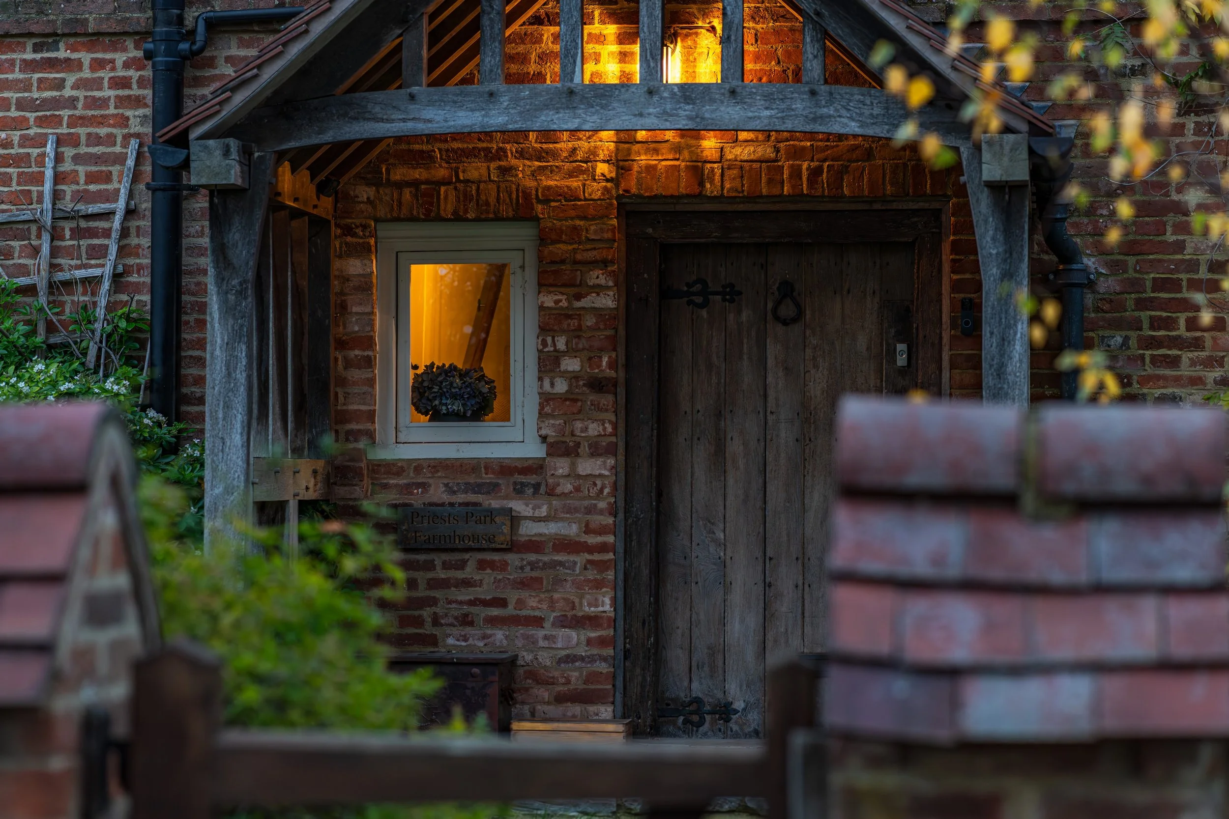 The entrance of a rustic brick farmhouse with a wooden door and small window, illuminated by warm interior lighting, surrounded by greenery and outdoor garden features.