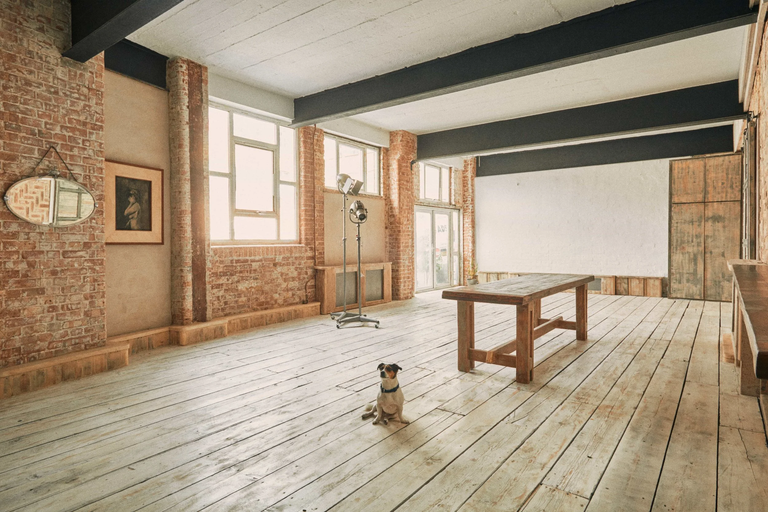 Interior of a spacious, industrial-style room with exposed brick walls, large windows, a wooden floor, and a few decorative items. There is a small dog sitting on the floor and a wooden table in the center.