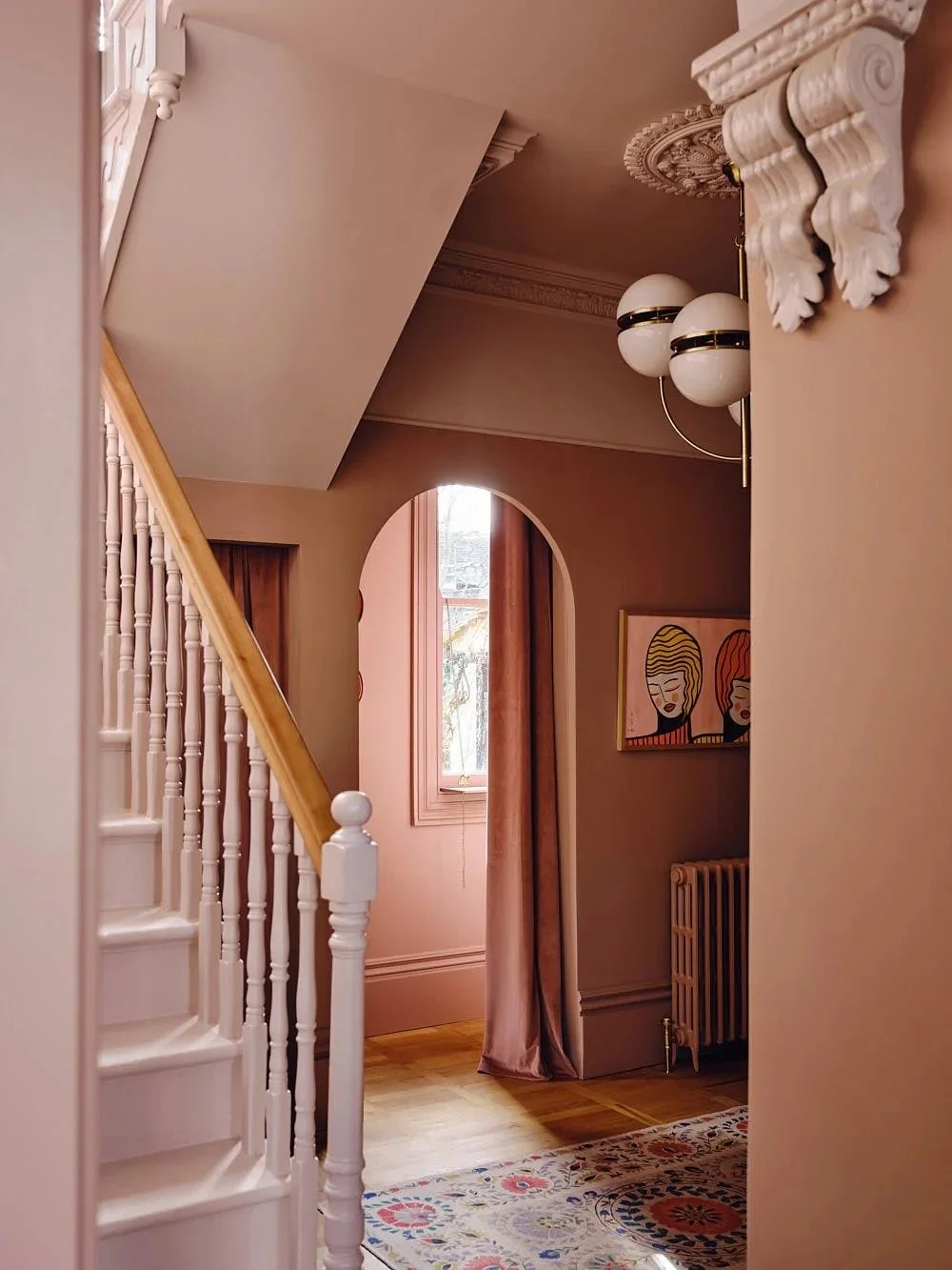 Interior view of a vintage-style room with a staircase, a window with curtains, a modern art painting, a radiator, a floral area rug, and decorative ceiling moldings.