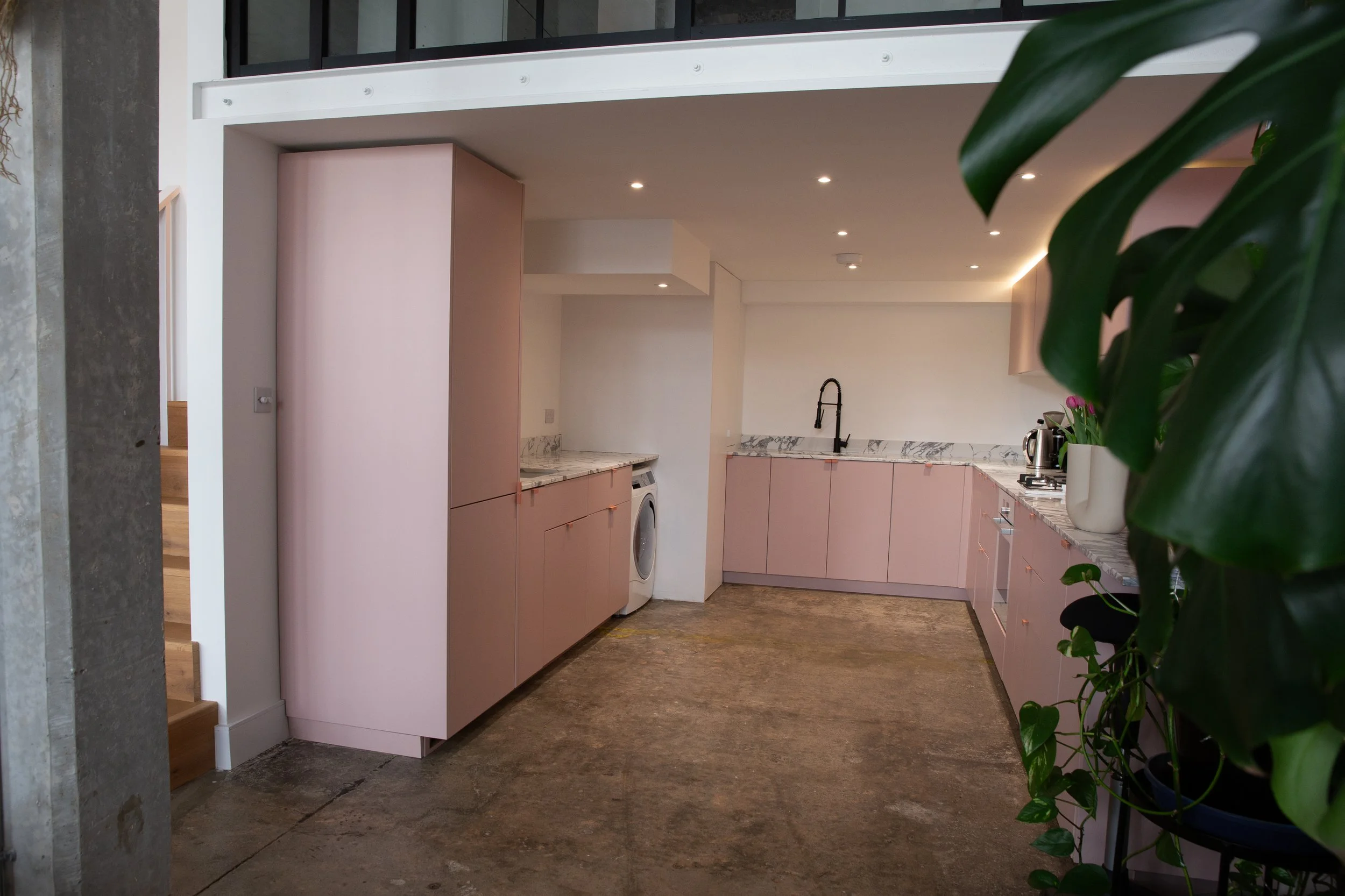 Modern kitchen with pink cabinets, marble countertops, black faucet, potted plant, and appliances, with an unfinished concrete floor.