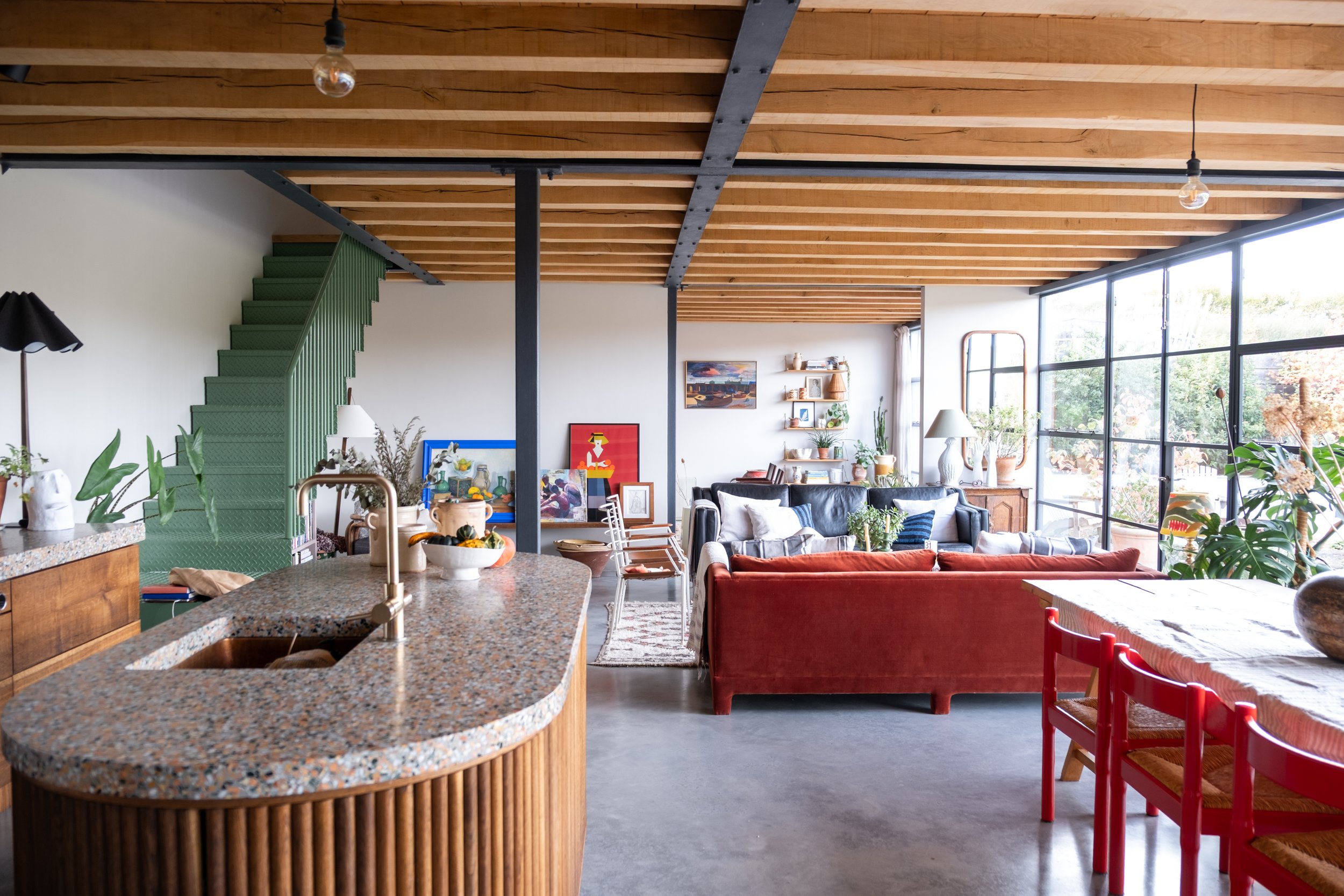 A spacious, modern living room with large floor-to-ceiling windows, wooden beam ceiling, and mixed furniture including a black leather sofa, red chairs, and various plants and art decorations.