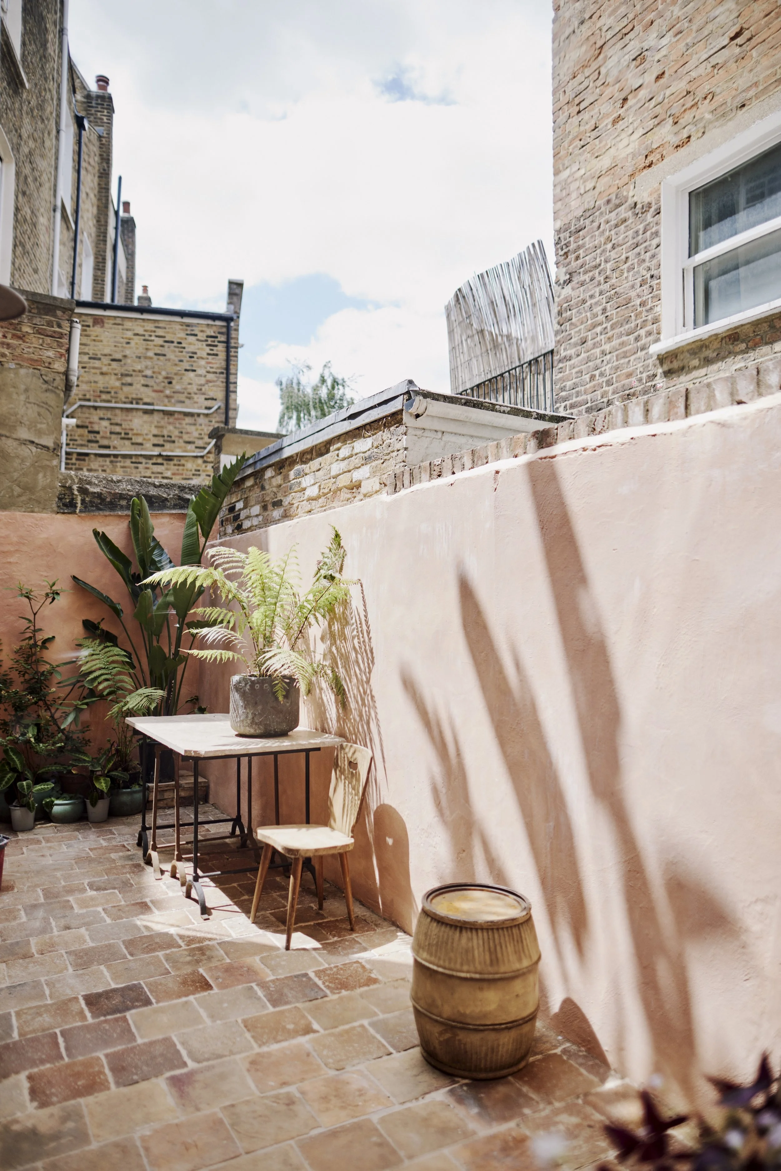 A small outdoor patio with a peach-colored wall, terracotta tile floor, and potted plants, including large green leaves and ferns. There is a white marble table with black metal legs, a small wooden chair, and a round wooden barrel, with sunlight cas