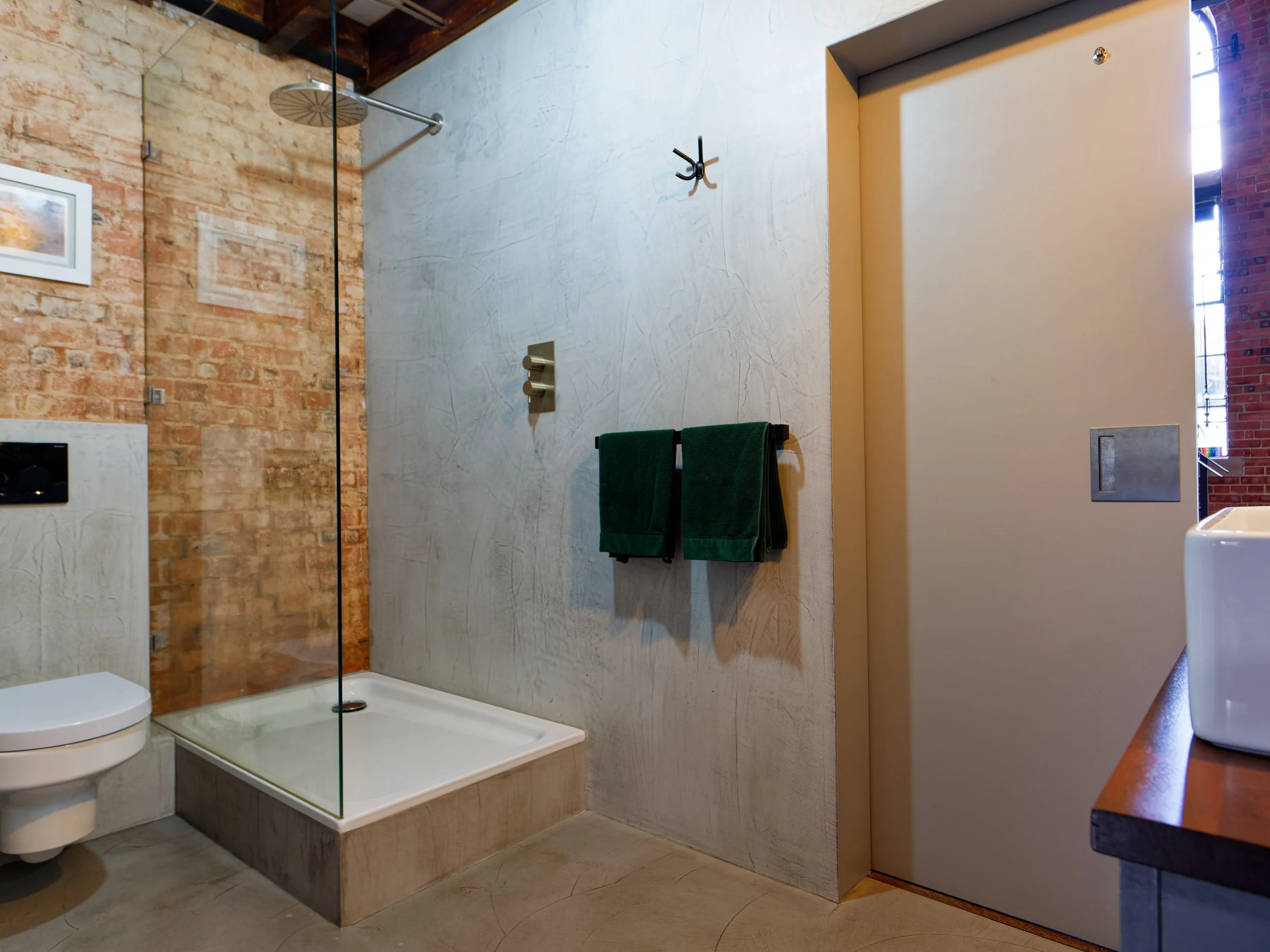 Modern bathroom with brick wall and concrete walls, walk-in shower with glass door, toilet, green towels, and a wooden countertop with a white container.