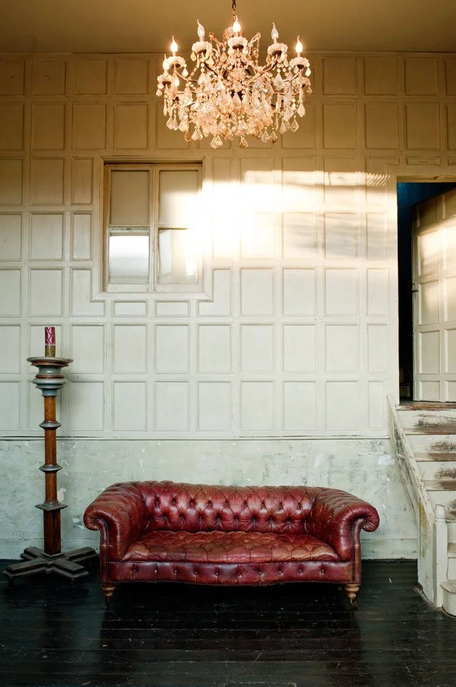 Elegant chandelier hanging from ceiling, red leather tufted sofa on dark hardwood floor, wall panels, small window, staircase on the right in a vintage-style room with aged walls.