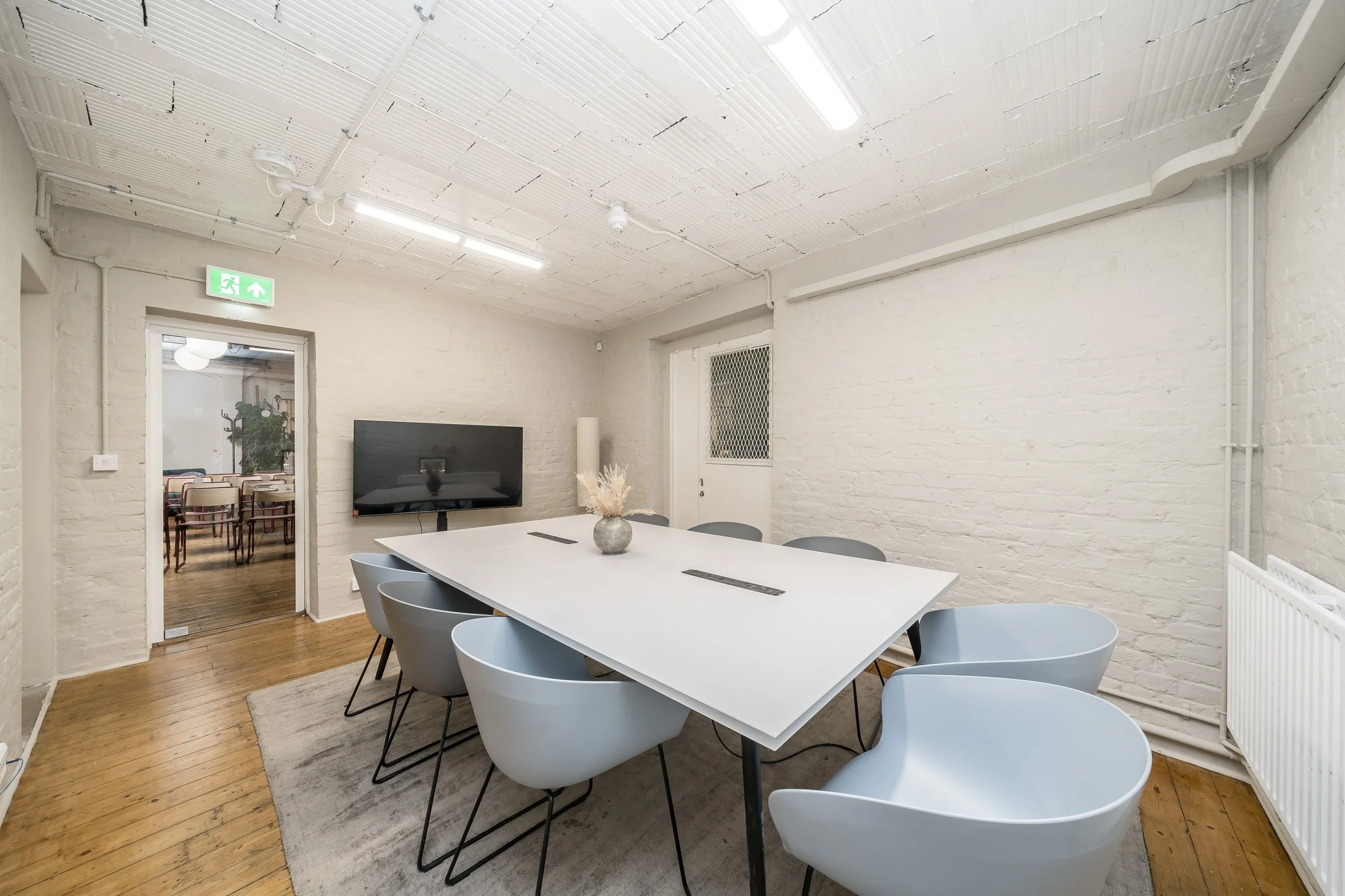 A modern conference room with white brick walls, a large white table, eight gray chairs, a wall-mounted television, a vase with dried flowers, and a wooden floor. There is an exit sign above an adjacent room.