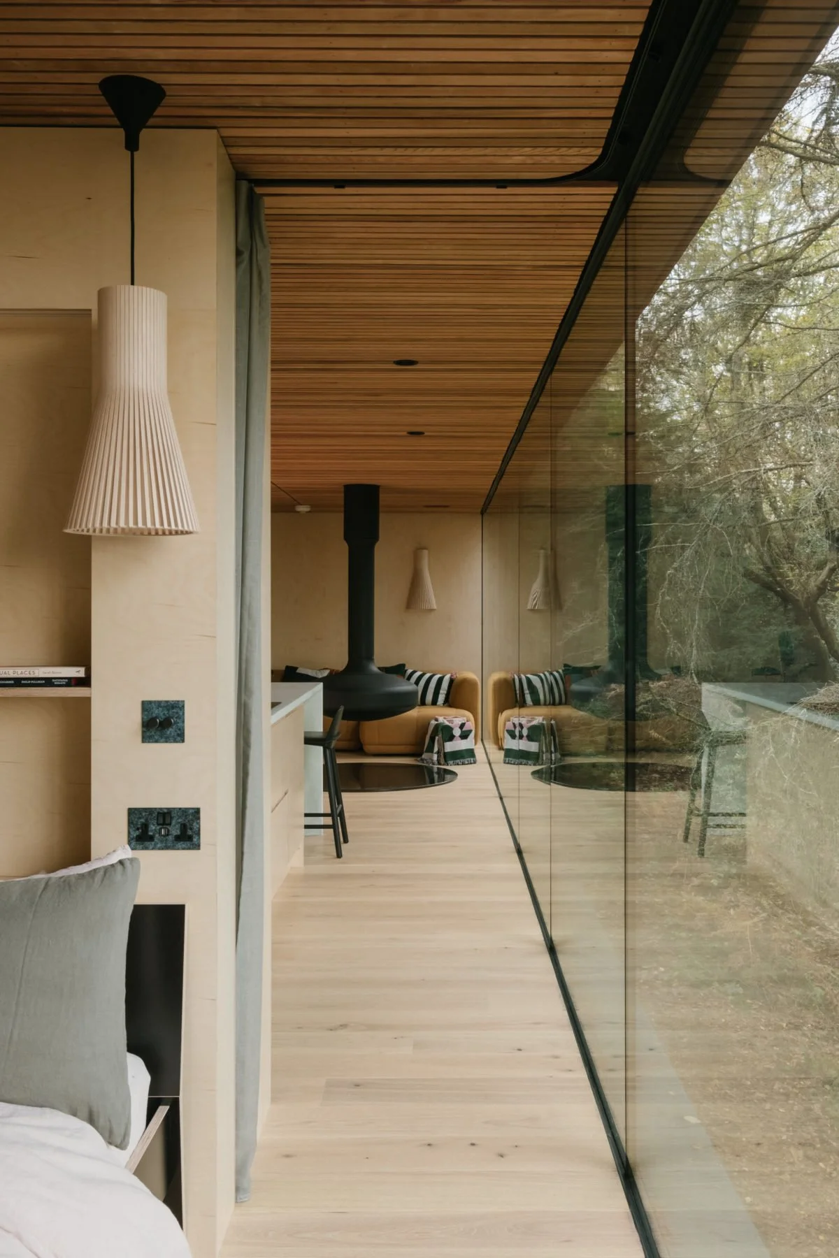 Modern interior living space with wooden ceiling, beige walls, a black chimney in the corner, a large glass window reflecting trees outside, and seating with striped and patterned pillows.