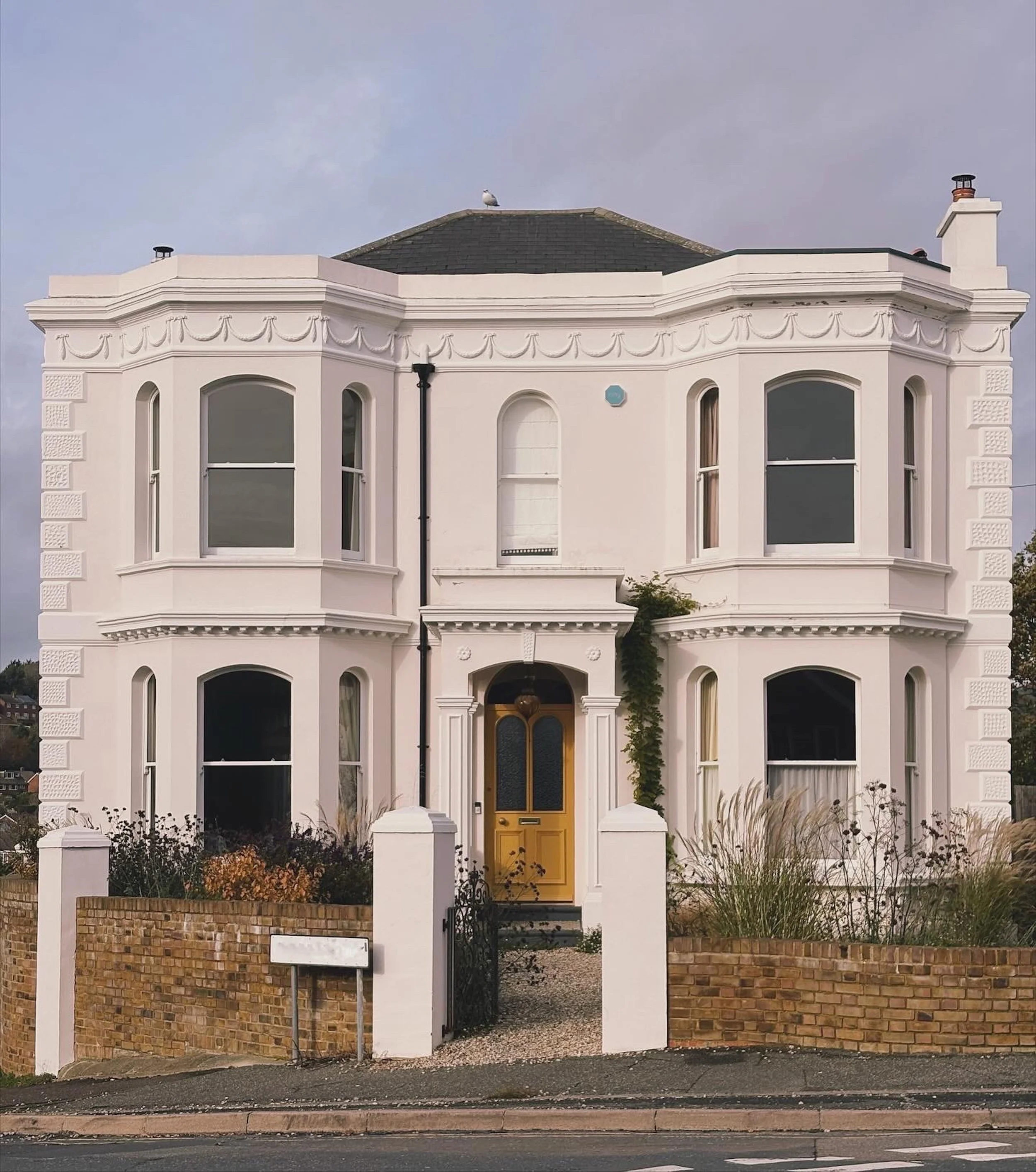 A three-story white Victorian house with a yellow front door, large bay windows on each floor, decorative trim, a brick wall with a gate at the entrance, and some greenery in the front yard.