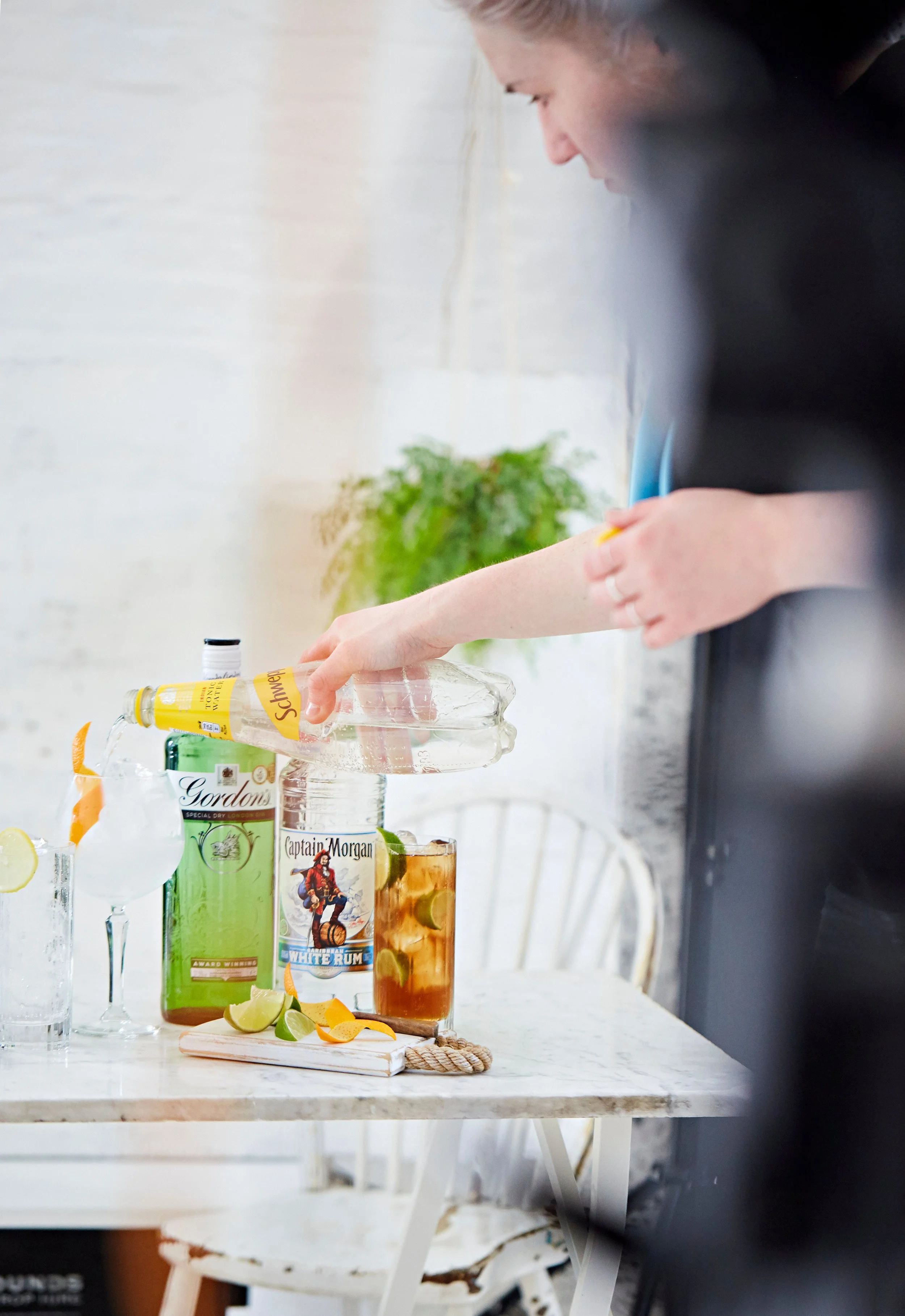 Person pouring a beverage into a glass at a bar or counter with bottles of alcohol and garnishes.
