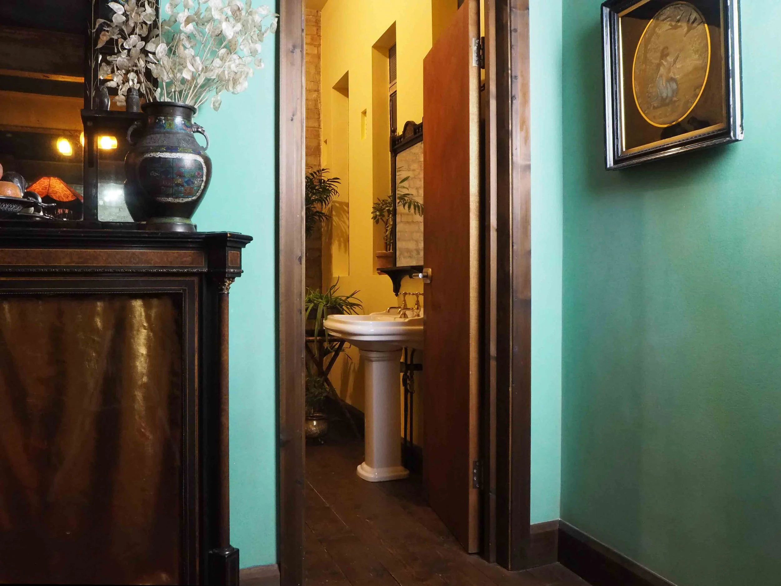 Part of a cozy indoor space with a turquoise wall on the right and a dark wooden cabinet on the left holding a black ceramic vase with white dried leaves. In the background is a bathroom with a white pedestal sink, plants, and glowing yellow walls. A