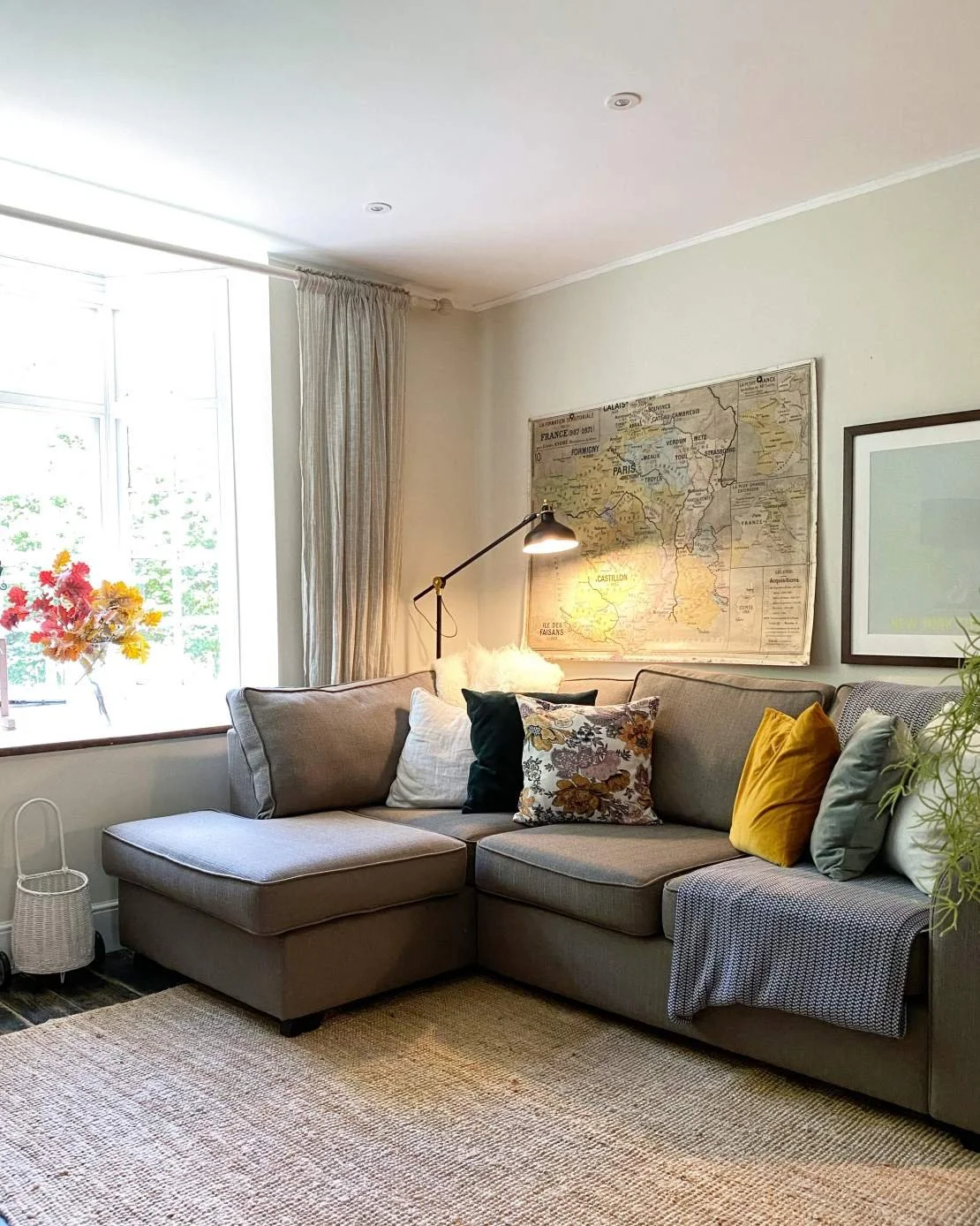 A living room with a gray sectional sofa, decorated with various colored pillows, a window with curtains, a framed map on the wall, and a Lamp on the corner