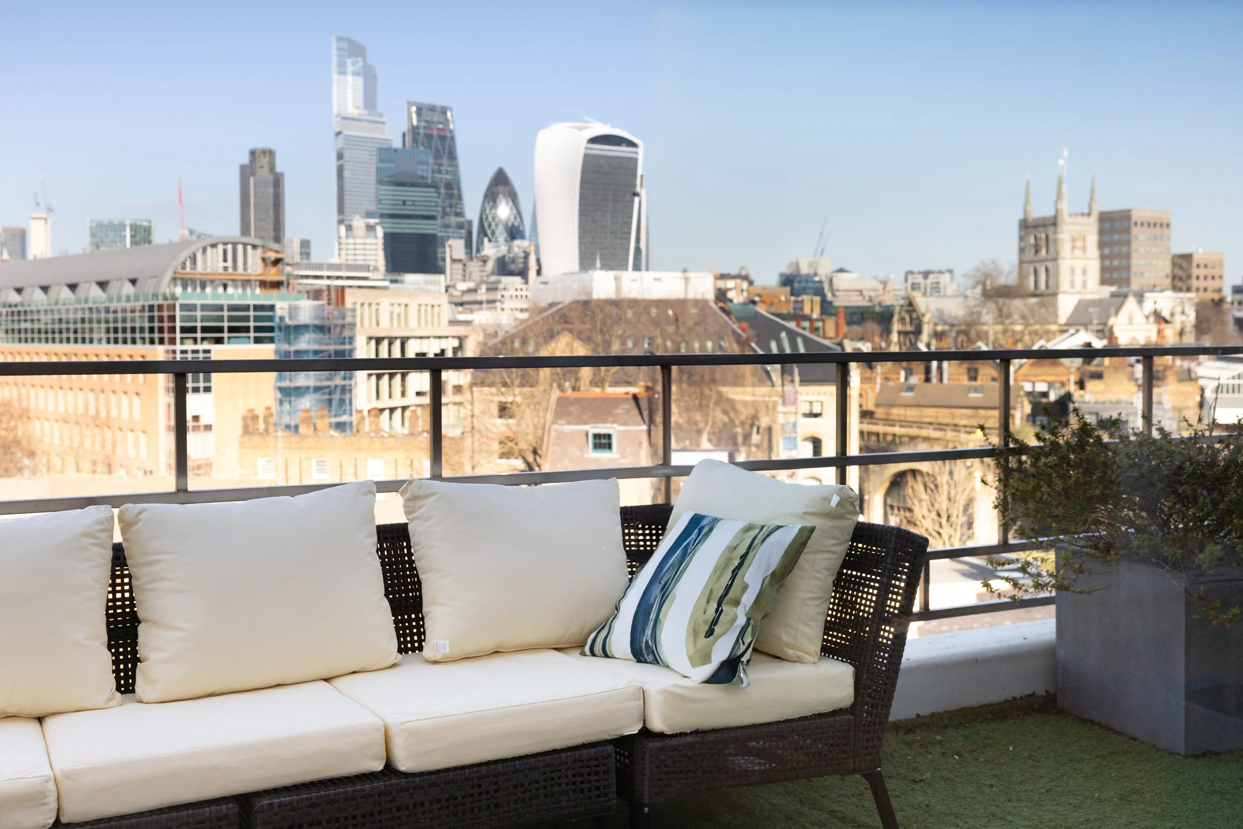 Outdoor balcony with beige cushioned sofa and decorative pillow, overlooking a city skyline with skyscrapers and historic buildings under a clear blue sky.
