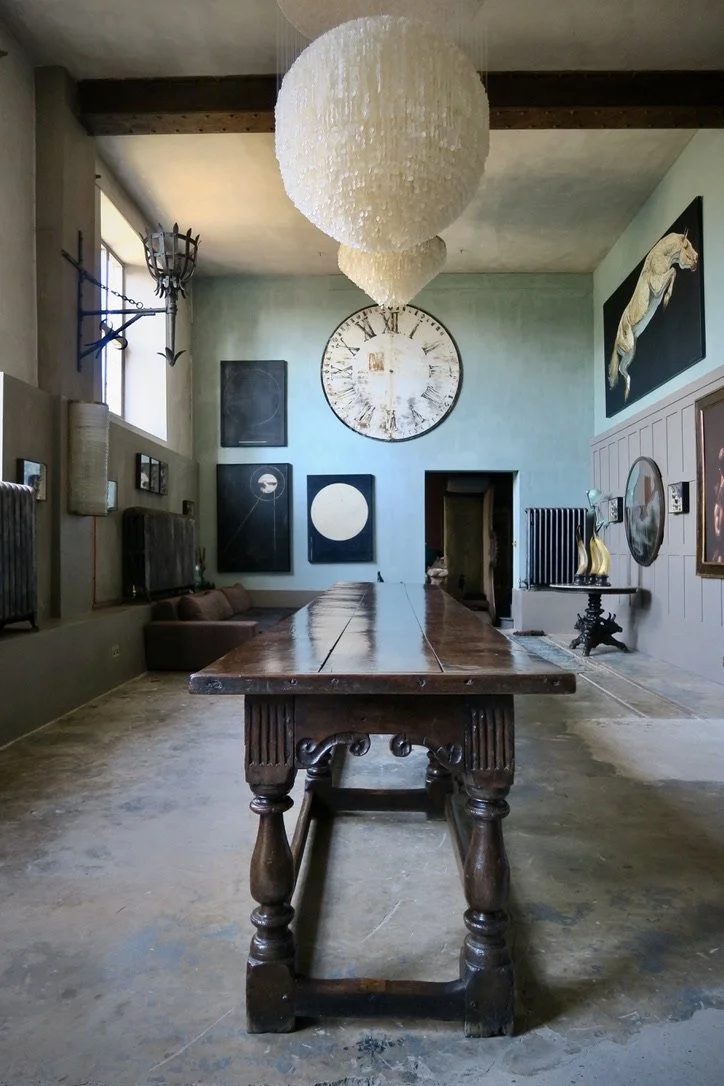 A rustic dining or conference room with a large wooden table, hanging pendant lights, a wall clock, and artwork including a rearing horse, framed pictures, and abstract pieces.