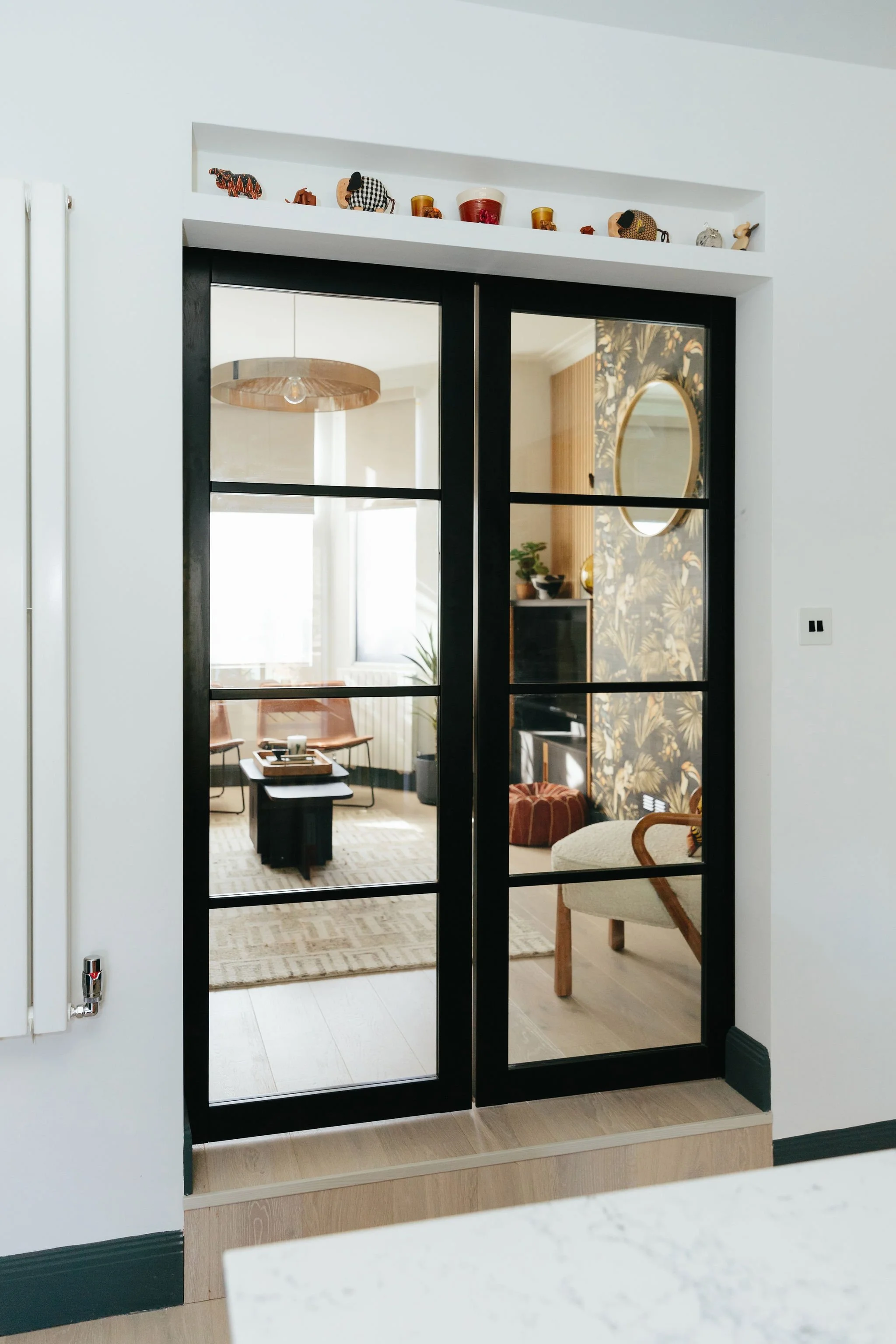 Glass sliding door with black frame leading to a living room with wooden flooring, a rug, chairs, a coffee table, a patterned wallpapered wall, a mirror, and decorative items.