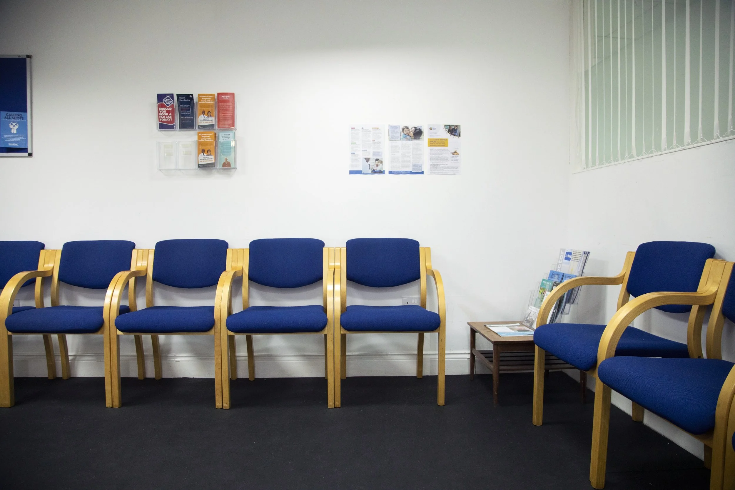 Empty waiting room with blue chairs and wooden armrests, some chairs have brochures and magazines, white wall with posters and informational materials, window with vertical blinds.