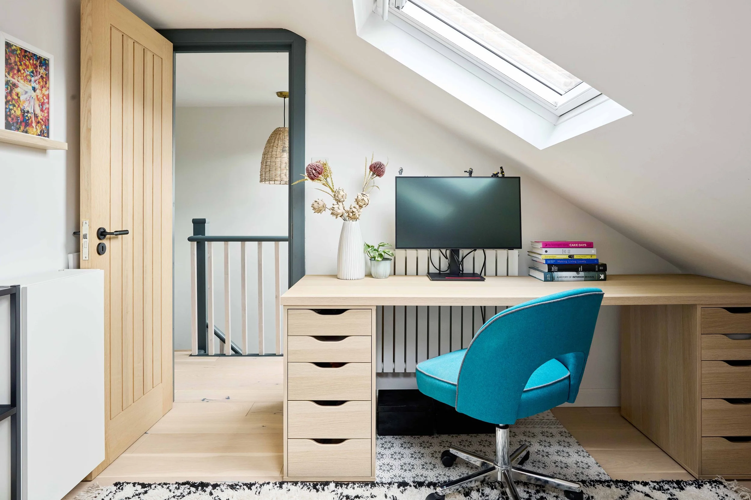 Home office with a desk, blue chair, computer monitor, stacked books, vase with dried flowers, and a skylight window in the sloped ceiling.