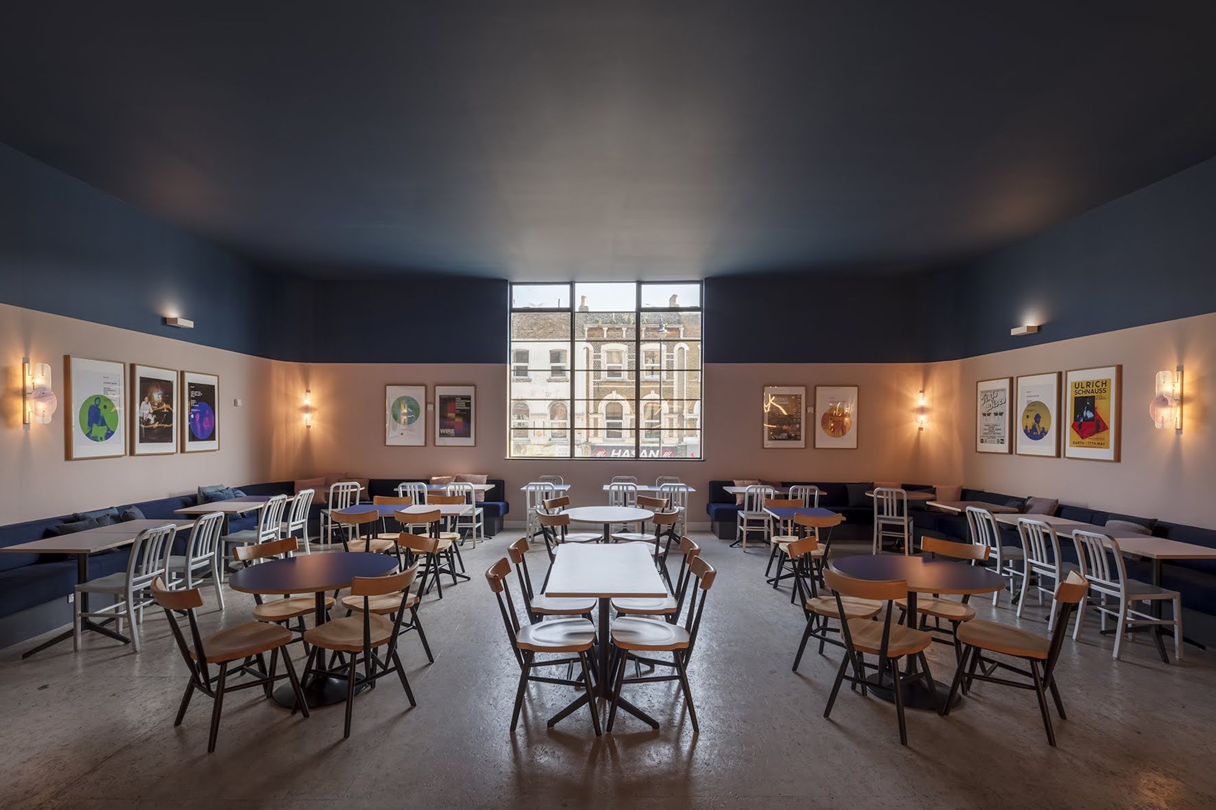 Empty modern restaurant with white and wooden chairs around tables, blue walls with framed posters, large window showing buildings outside, soft wall lighting.