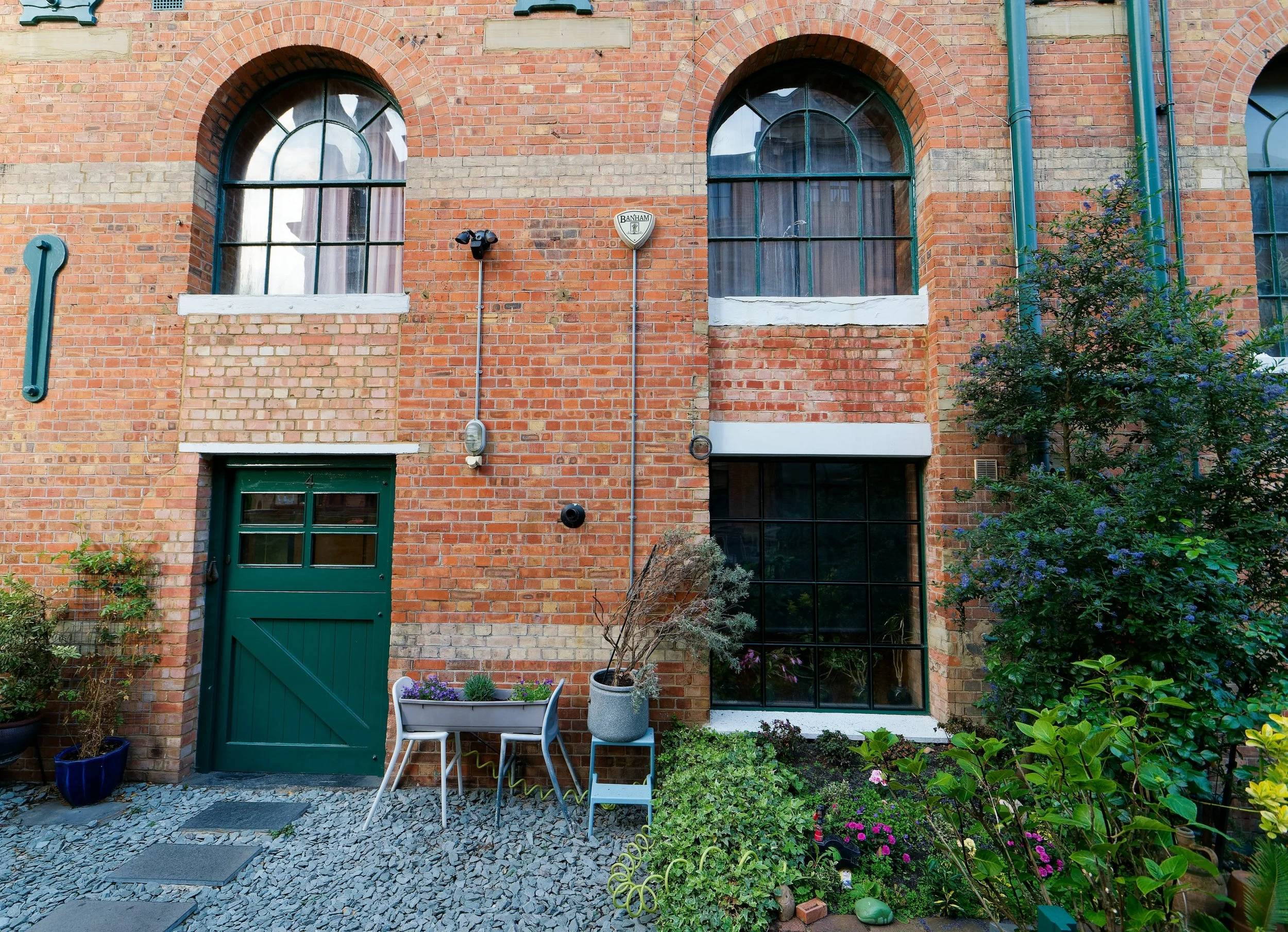 View of a brick building with arched windows, a green door, and outdoor plants and furniture in a small garden courtyard.