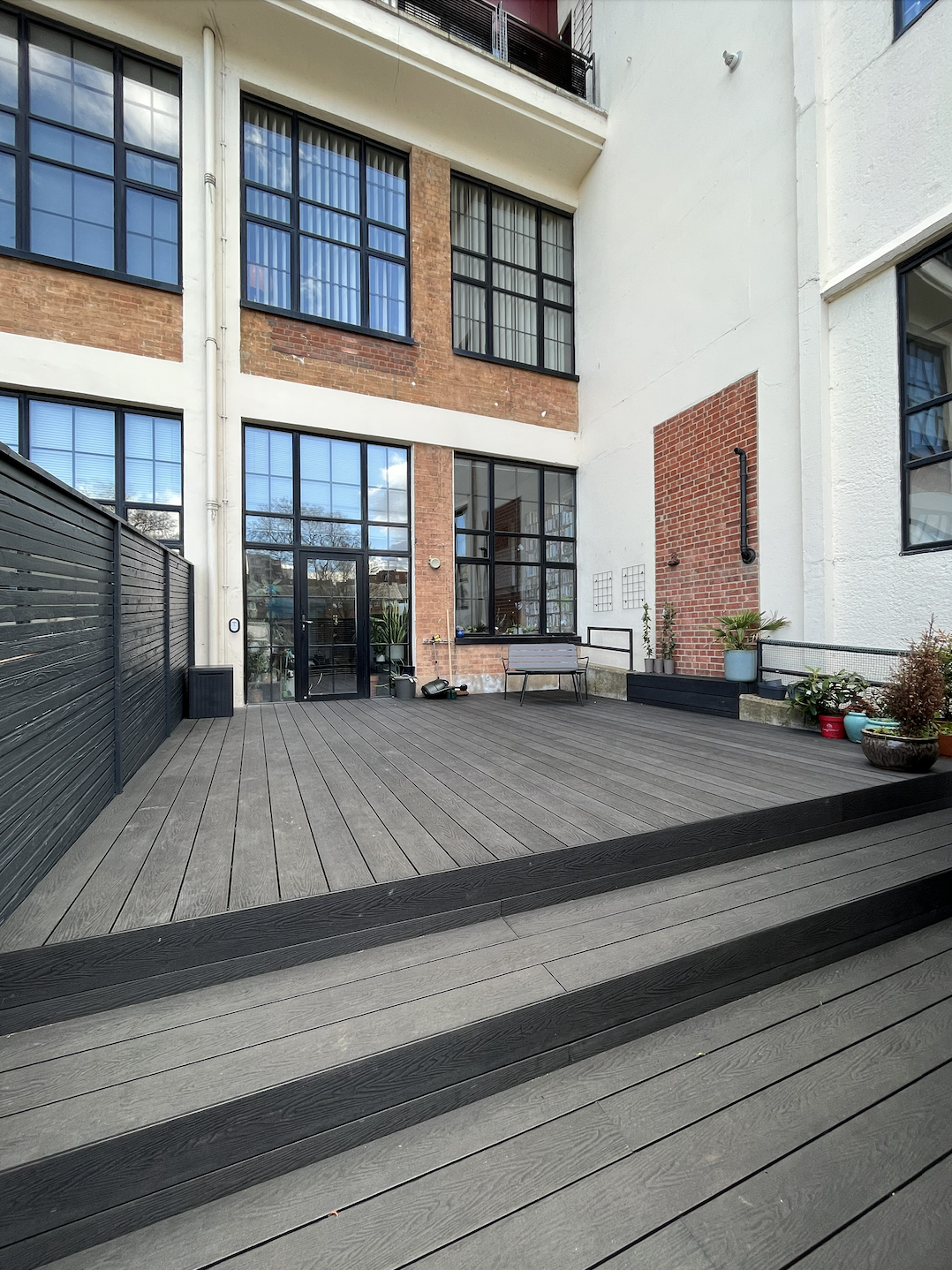 Urban outdoor patio with wooden flooring, planters, and glass doors leading to indoor space, surrounded by a modern multi-story building with large windows and brick accents.