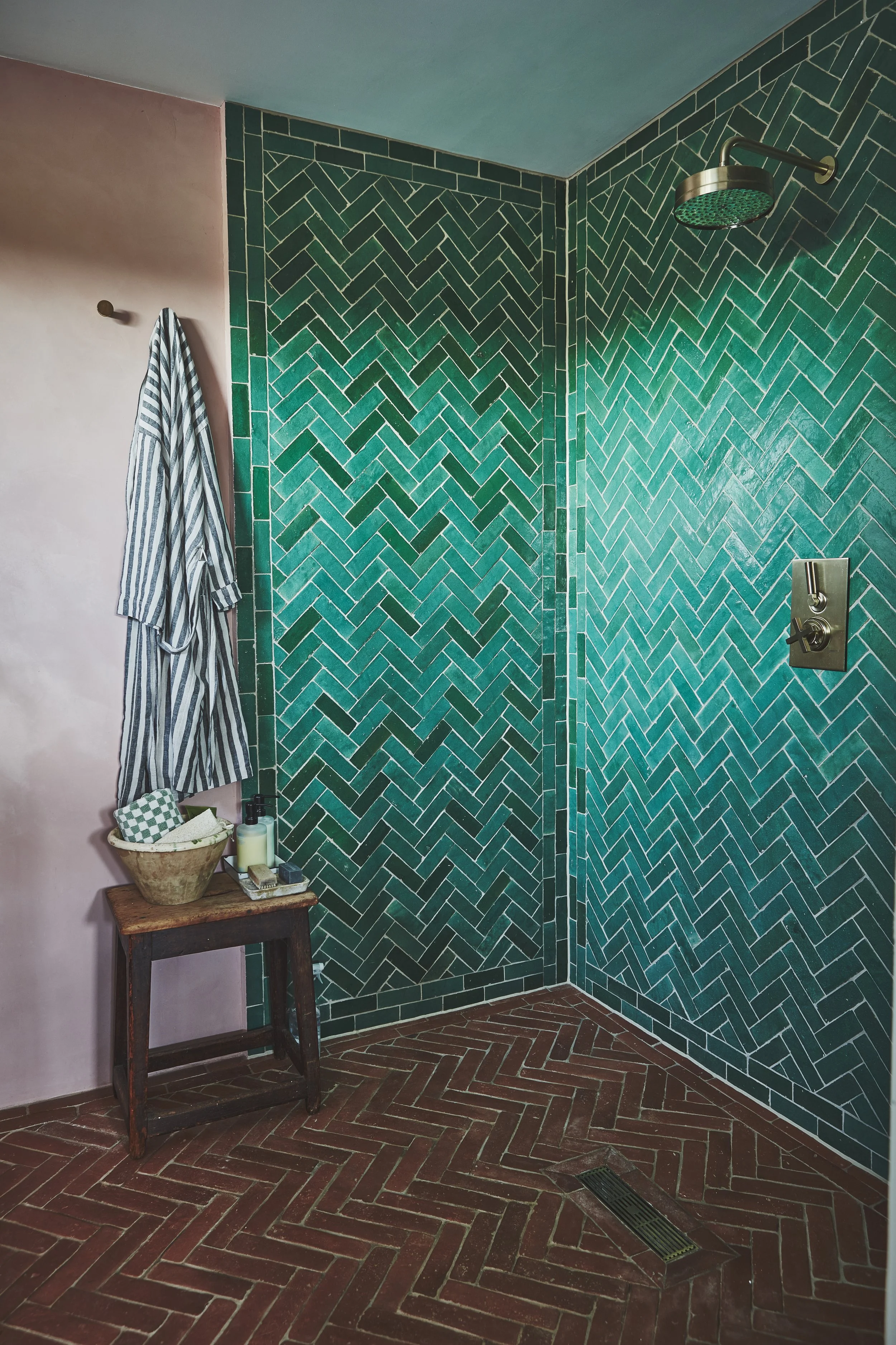 A bathroom shower area with green herringbone tiles and a silver showerhead. A pink wall has a striped towel hanging on a wooden peg. Below the towel, a small wooden table holds a basket containing a towel and toiletry items. The floor is made of red