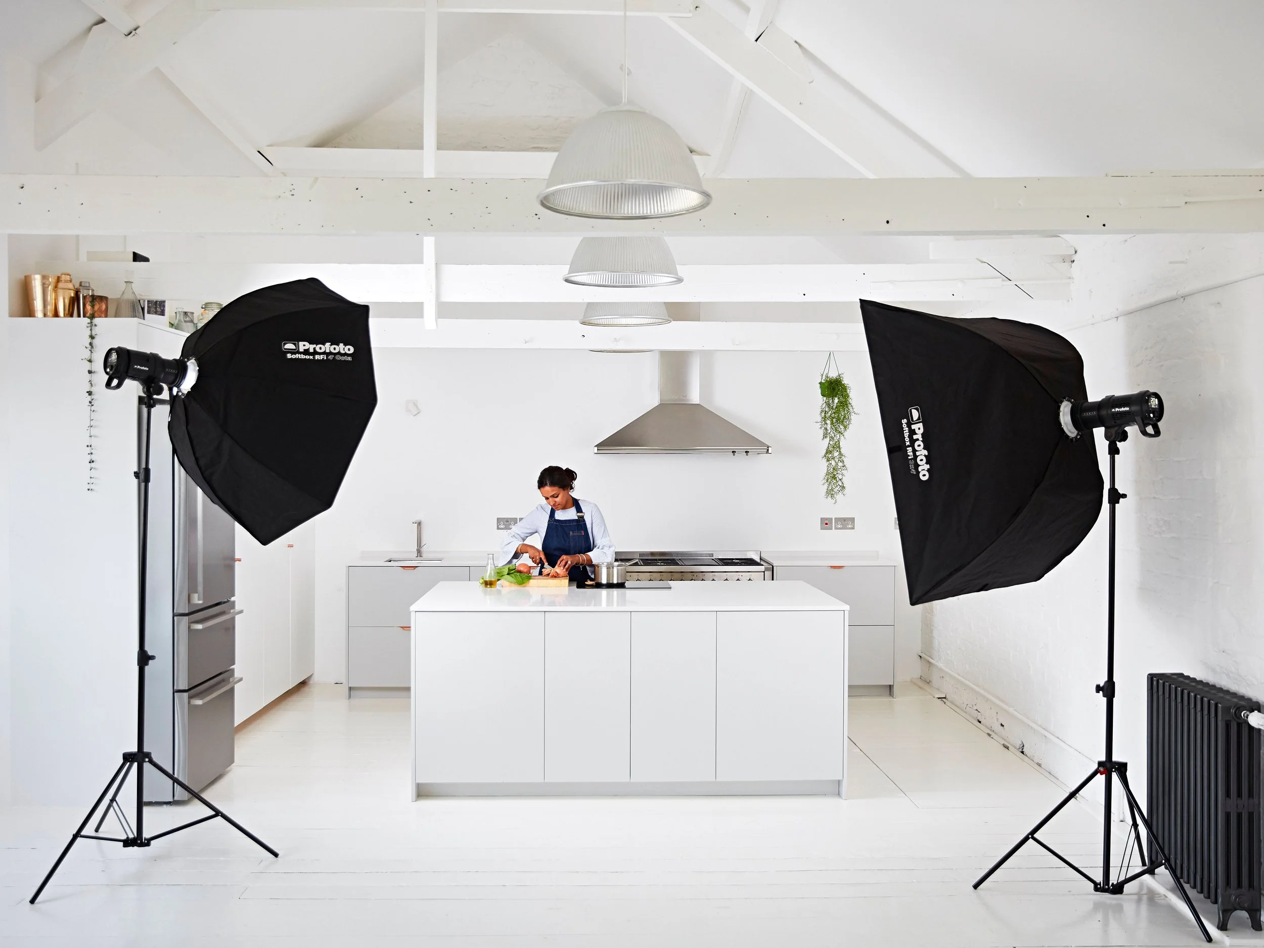 A woman cooking in a modern white kitchen studio with professional photography lighting equipment, including two large softbox lights on stands, and green plants hanging from the ceiling.