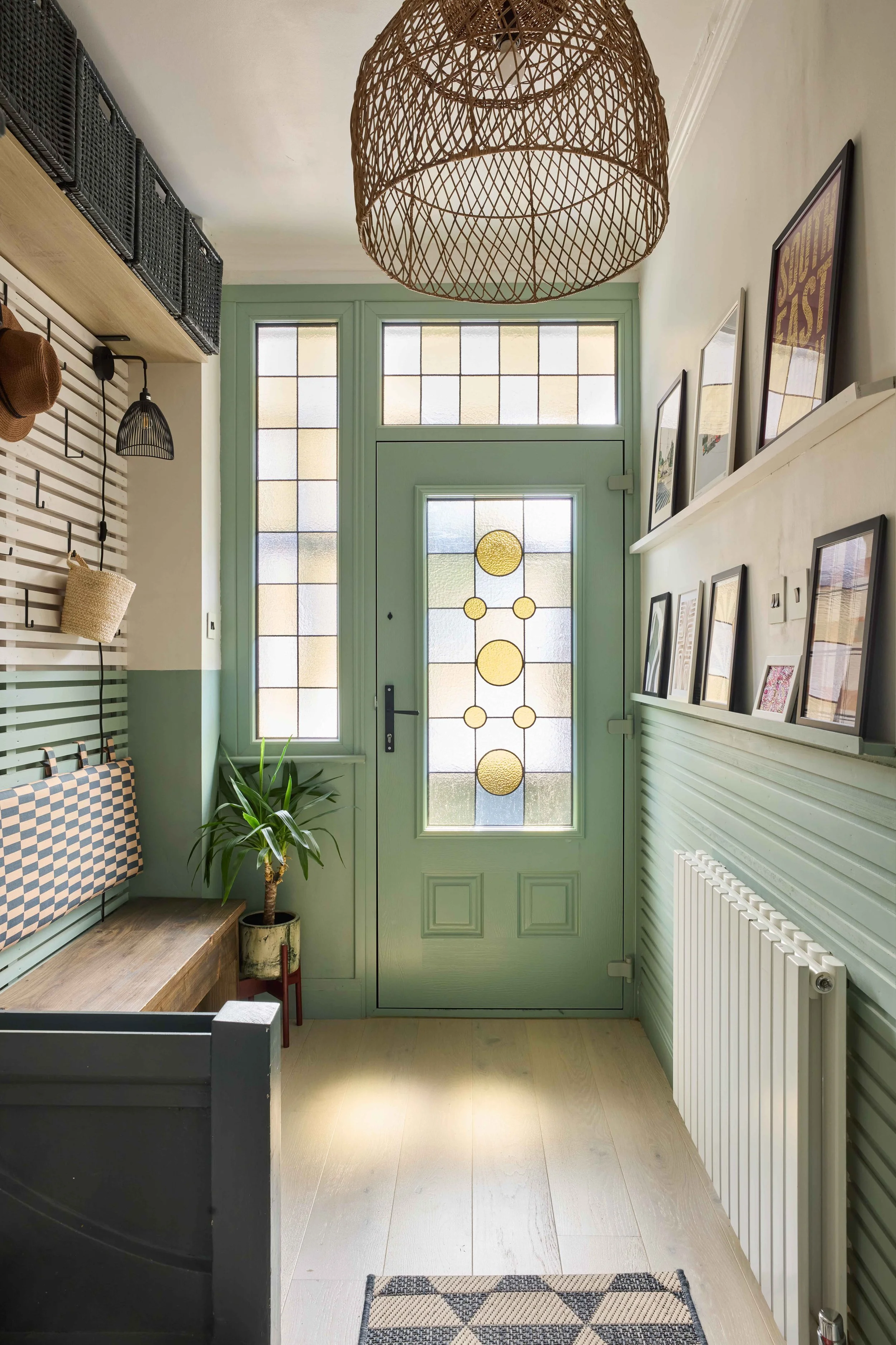 Entryway of a home with a green door featuring decorative glass, framed photos on the wall, a plant near the door, and a wicker pendant light hanging from the ceiling.