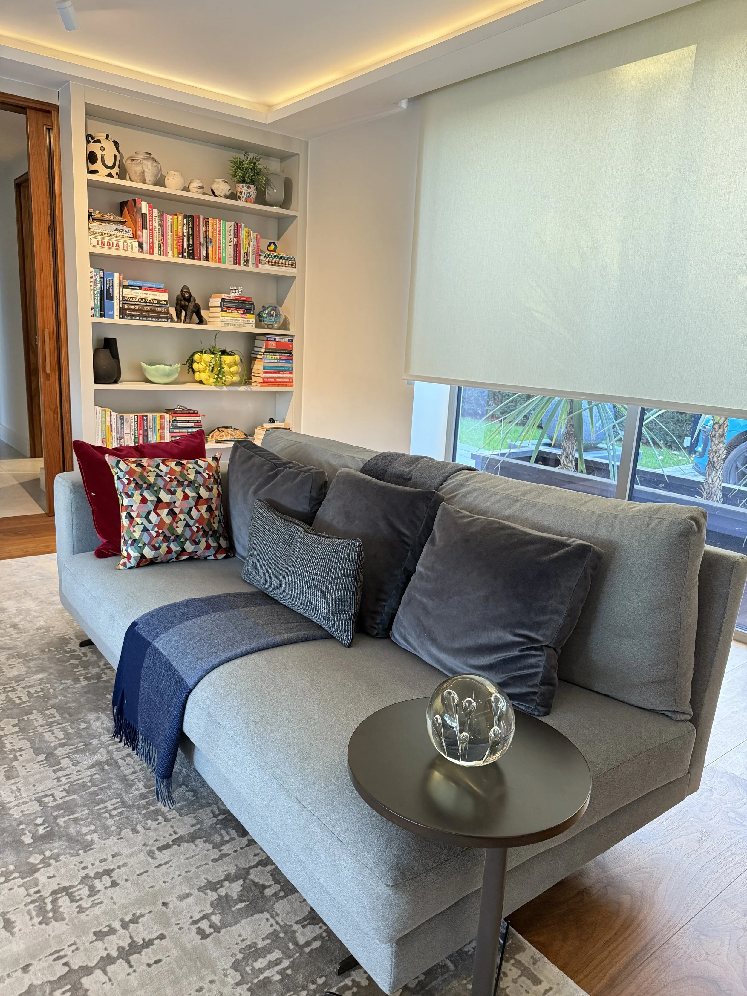Living room with a gray sofa decorated with patterned and dark solid-colored pillows, a side table with a glass ornament, a bookshelf filled with books and decorative objects, and a large window with a green shade, revealing a view outside.