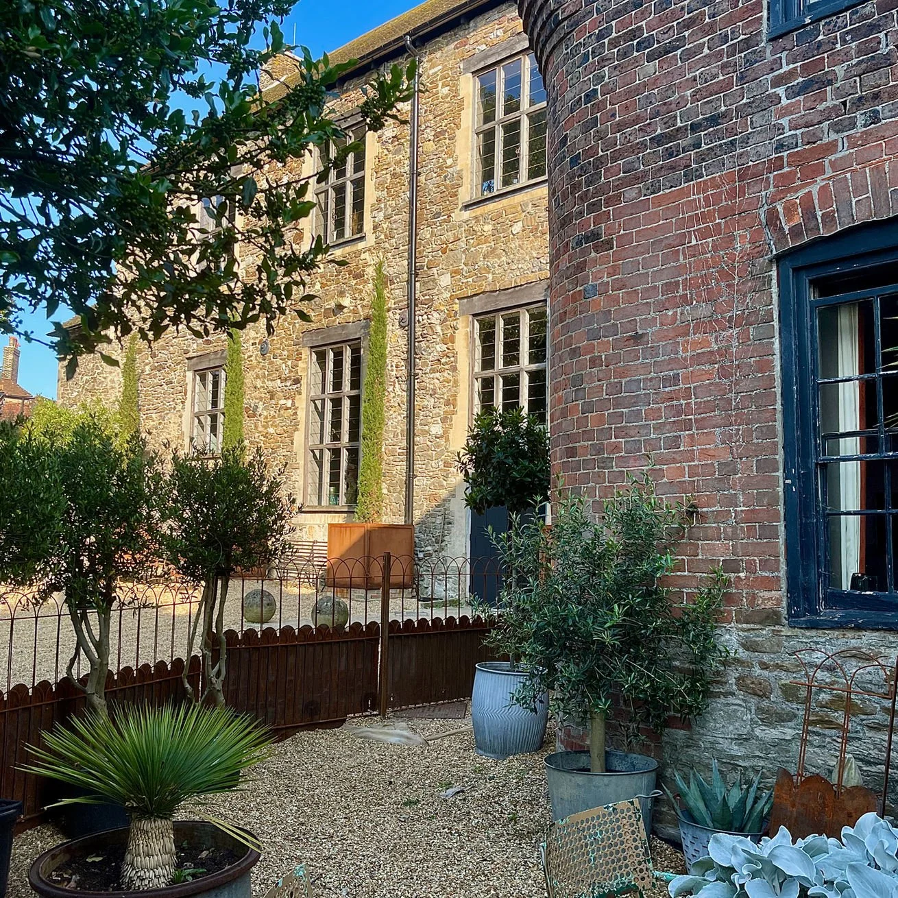 A small garden with potted plants, including a palm and an agave, in front of a brick house with multiple windows and a stone wall. A wood fence separates the garden from a stone-paved area.